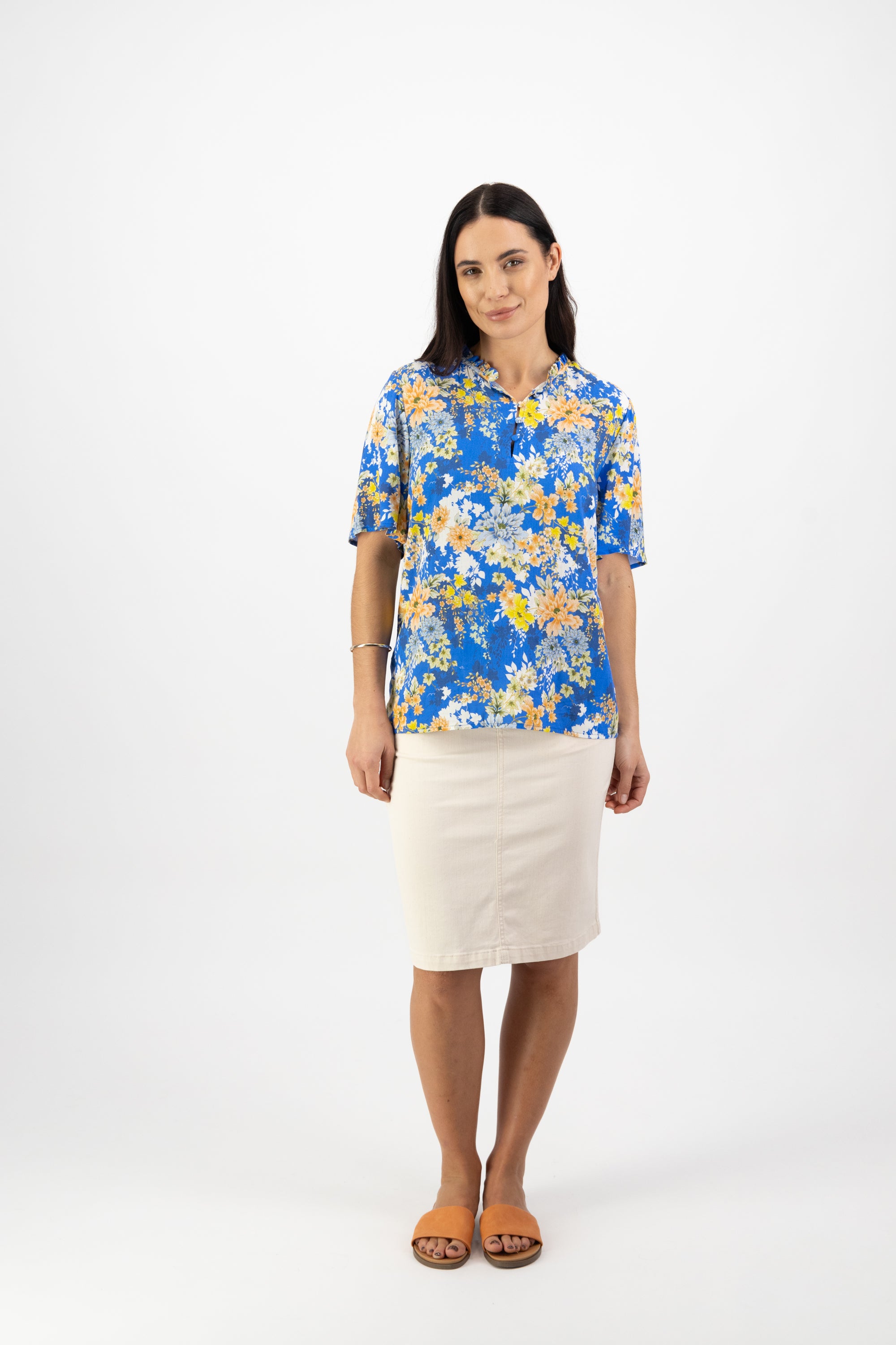 A woman with long dark hair and a neutral expression stands against a white background, wearing the Vassalli Floaty Sleeve Top - Kimono Dream in blue and yellow floral, a light beige knee-length skirt, and brown sandals.