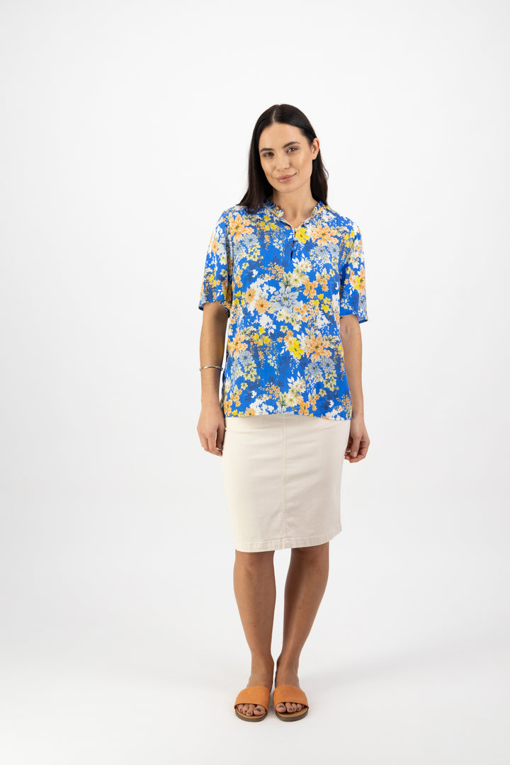 A woman with long dark hair and a neutral expression stands against a white background, wearing the Vassalli Floaty Sleeve Top - Kimono Dream in blue and yellow floral, a light beige knee-length skirt, and brown sandals.