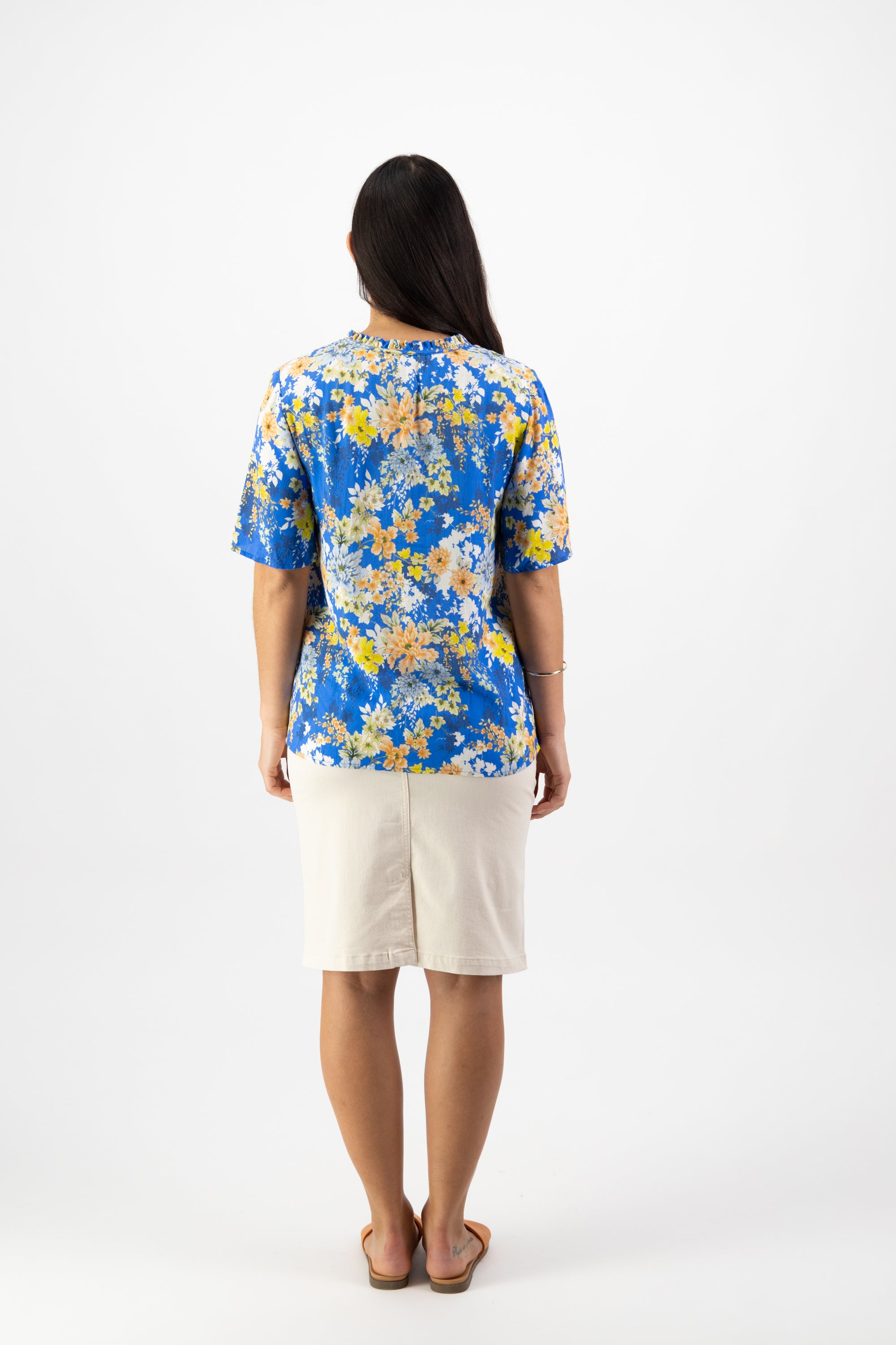 A woman stands facing away, wearing the Vassalli Floaty Sleeve Top - Kimono Dream in a blue, yellow, and white floral print, paired with a beige knee-length skirt and brown sandals against a plain white background.
