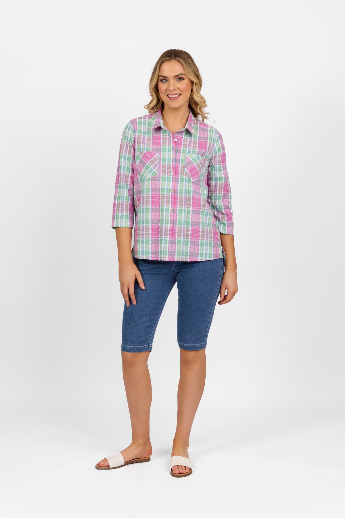 A woman smiles, wearing the Vassalli Button Front Shirt - Pink Check, paired with knee-length blue denim shorts and white slide sandals against a plain white background.