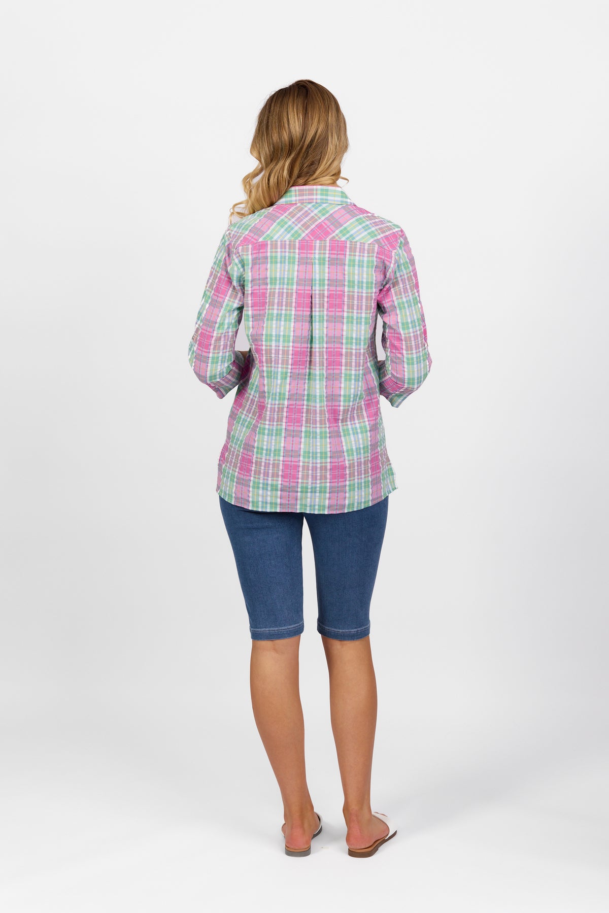 A woman faces away from the camera in a Vassalli Button Front Shirt - Pink Check, blue knee-length shorts, and flip-flops, set against a plain white background.