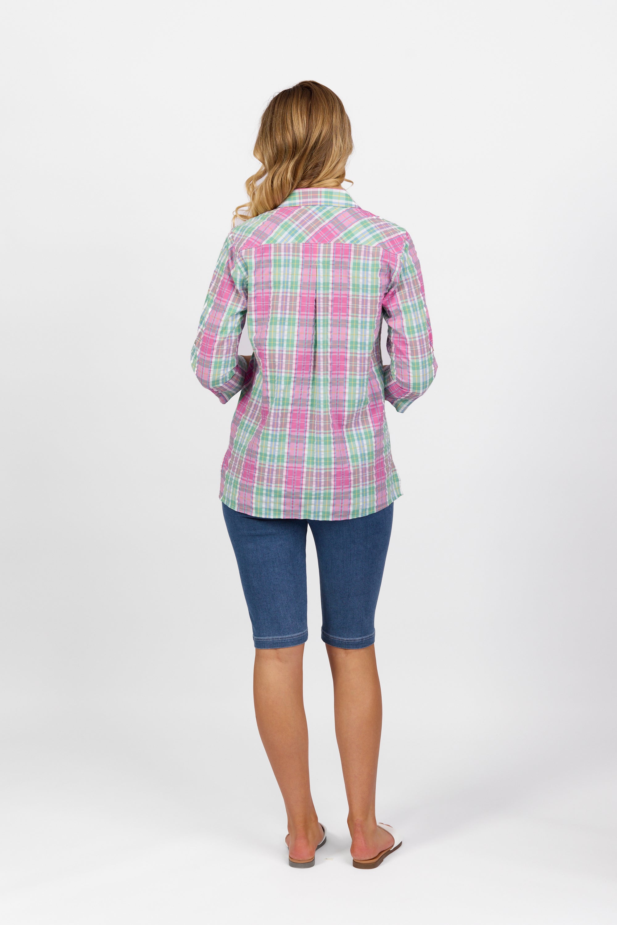 A woman faces away from the camera in a Vassalli Button Front Shirt - Pink Check, blue knee-length shorts, and flip-flops, set against a plain white background.