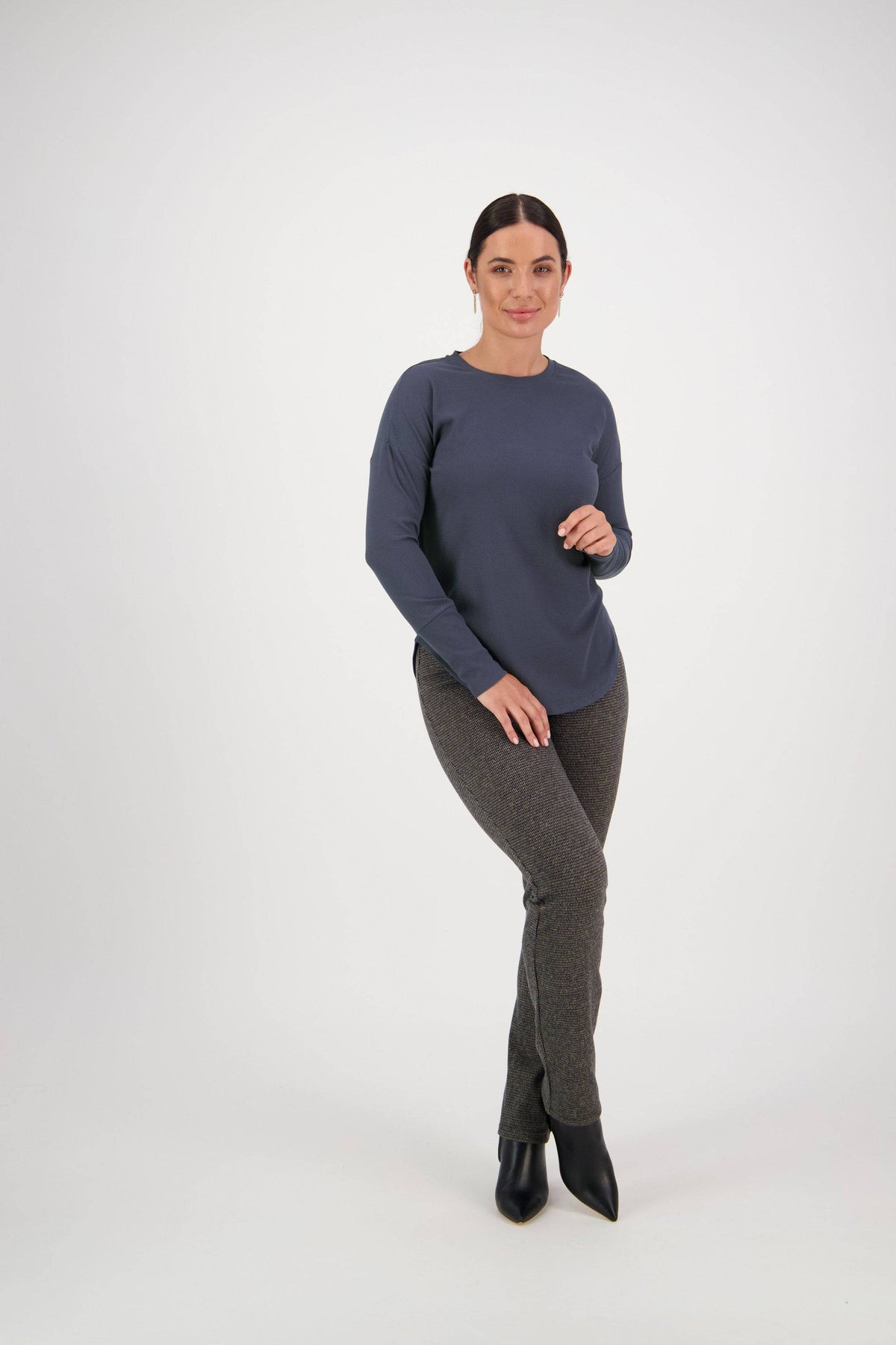 A woman with dark hair in a low bun poses against a white background, wearing the Vassalli Round Neck Top in denim blue, paired with gray pants and black ankle boots. She smiles slightly, standing with one knee bent.