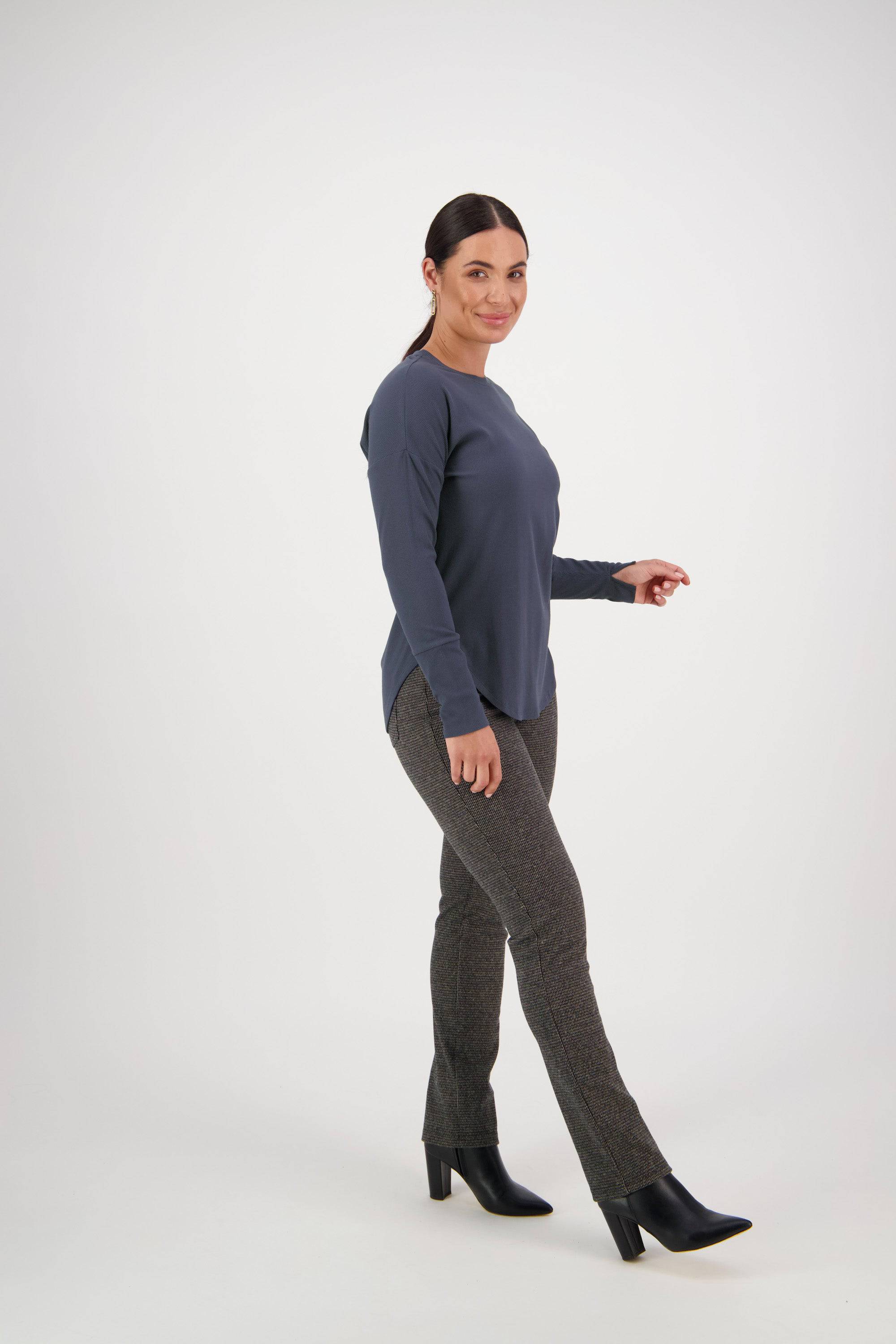 A woman with dark hair in a ponytail walks and smiles at the camera, wearing a Vassalli Round Neck Top in denim blue, gray straight-leg pants, and black heeled boots against a plain white background.