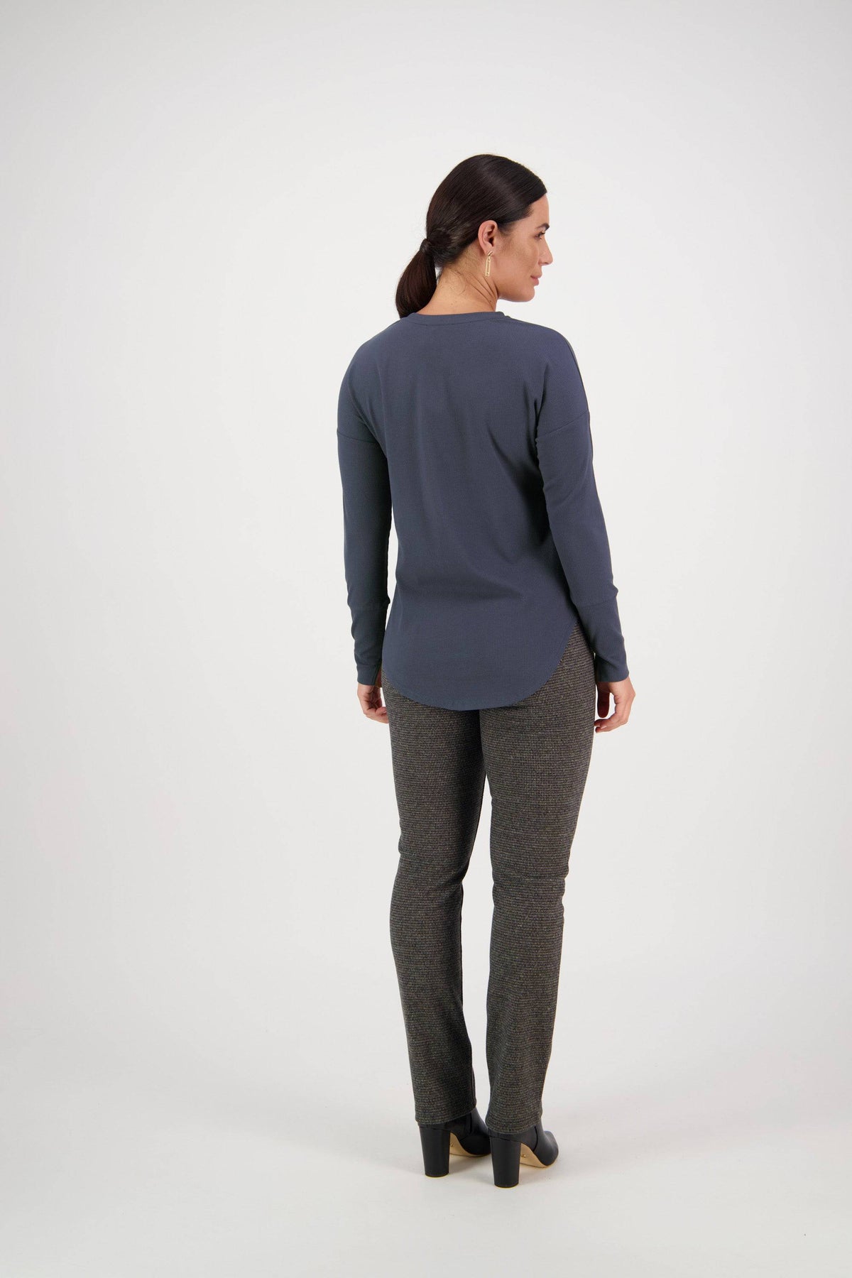 A woman with dark hair in a ponytail faces away, wearing the Vassalli Round Neck Top in denim blue, paired with gray pants and black heeled shoes, against a plain white background.