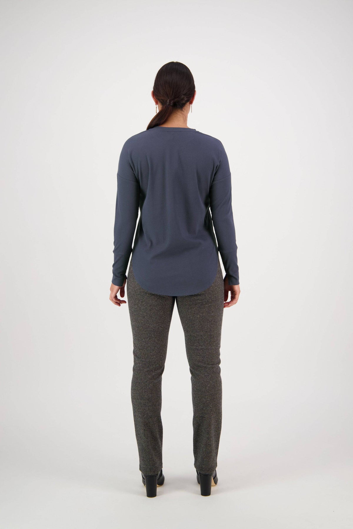 A person with long hair tied back models the Vassalli Round Neck Top in Denim Blue, paired with gray pants and black shoes, standing with back to camera against a plain white background.