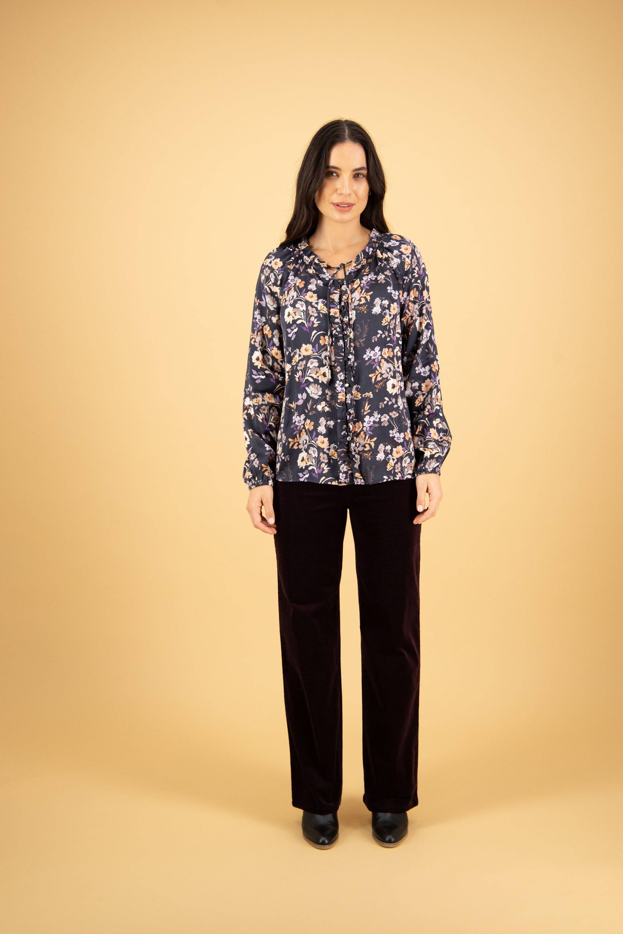 A woman with long dark hair poses against a beige background, wearing the Vassalli V-Neck Top - Flora in navy blue floral print, with dark wide-leg pants and black shoes. She stands facing forward with a neutral expression.