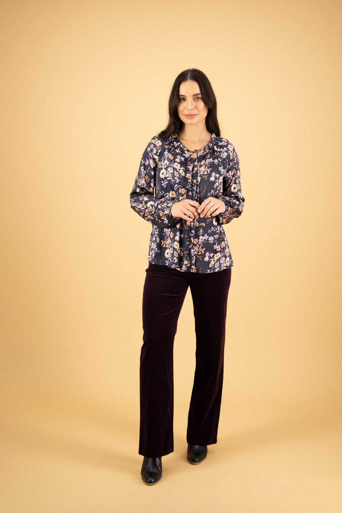 A woman with long dark hair stands against a beige background, wearing the Vassalli V-Neck Top - Flora, paired with dark velvet pants and black boots. She smiles gently, hands lightly clasped in front of her.