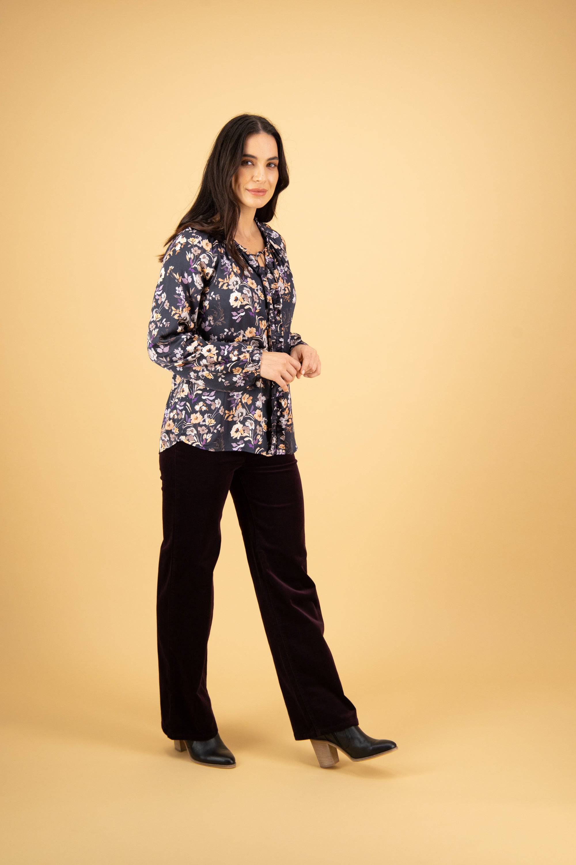 A woman with long dark hair stands against a beige background, wearing the Vassalli V-Neck Top - Flora, dark pants, and black ankle boots. She smiles slightly while facing the camera.