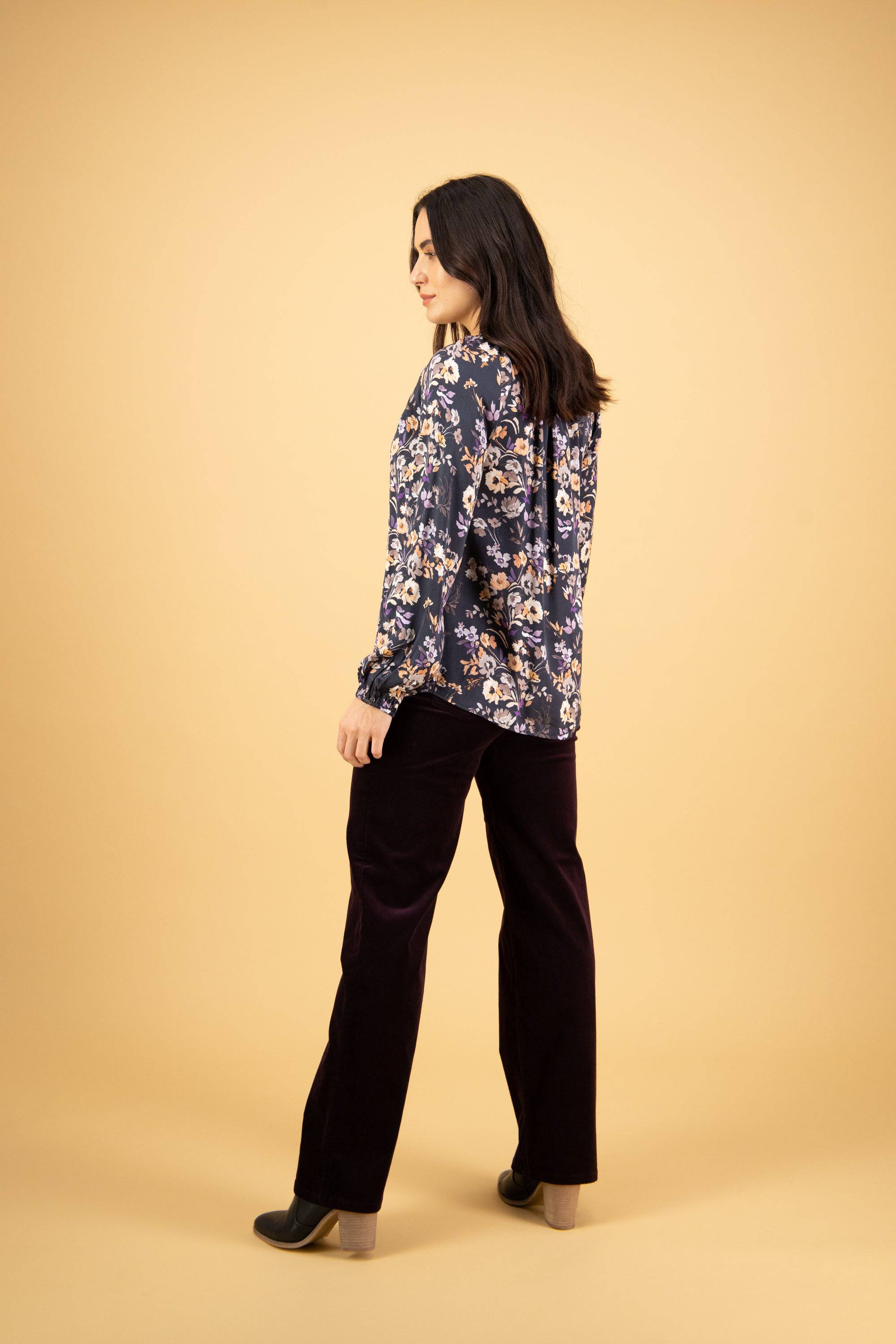 A woman stands against a beige background, wearing the Vassalli V-Neck Top - Flora with a navy floral print, paired with dark wide-leg pants and heeled shoes. She is turned slightly to the side, looking down with a relaxed expression.