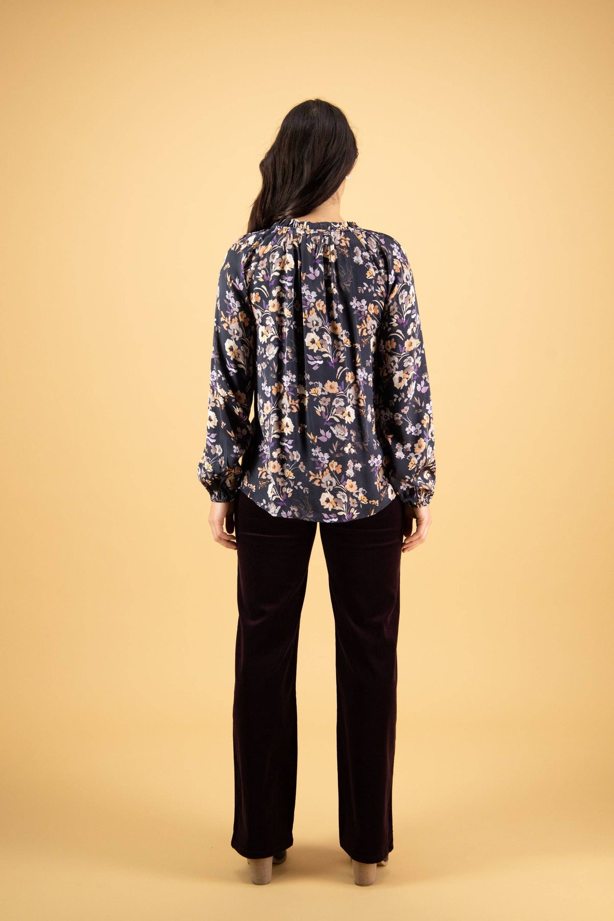 A woman with long dark hair stands facing away, wearing the Vassalli V-Neck Top - Flora in navy blue floral print and dark pants against a plain beige background.