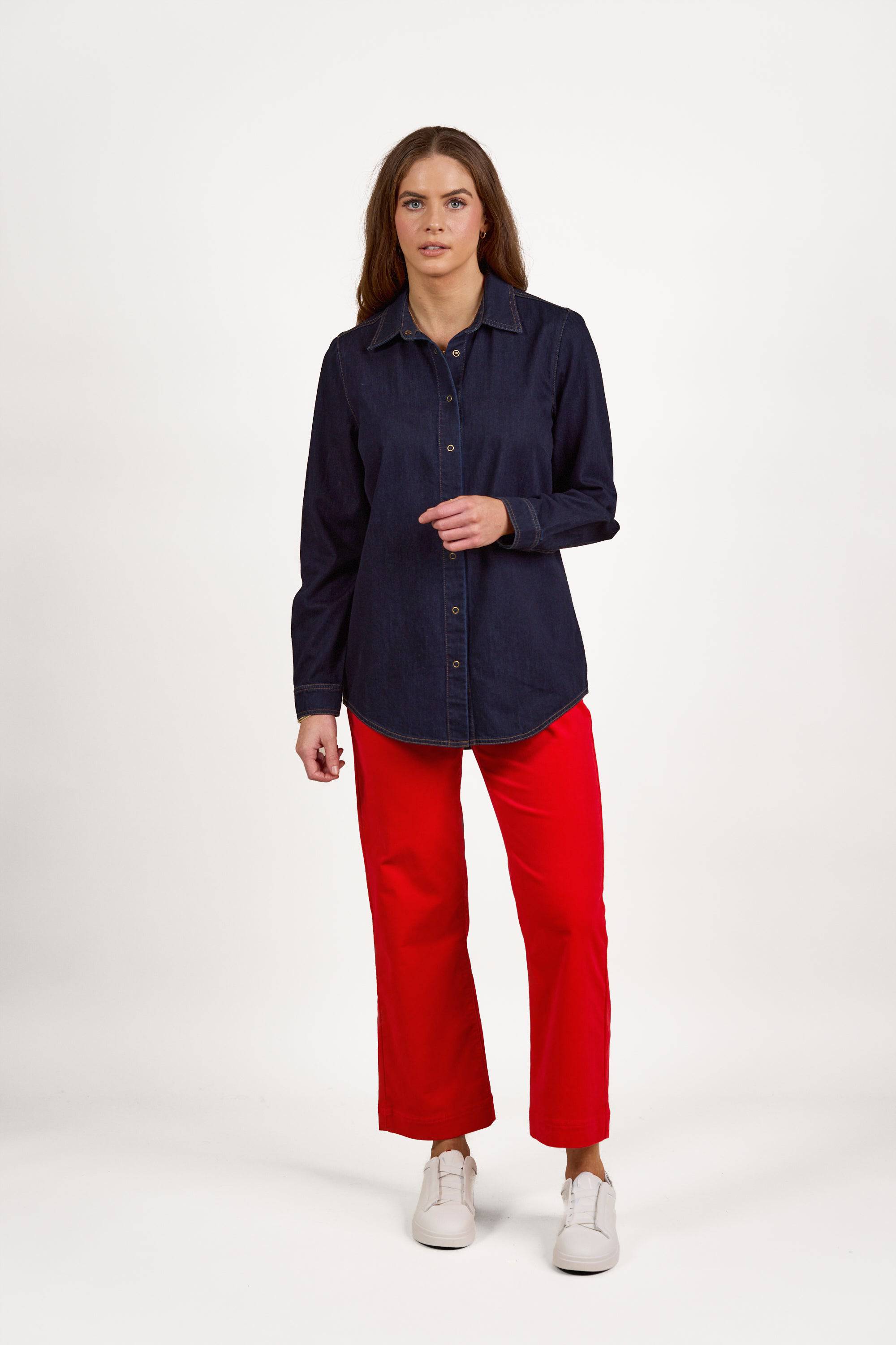 A woman with long brown hair wears the Vassalli Shirt With Poppers in Dark Denim, styled with bright red pants and white sneakers, standing against a plain white background.