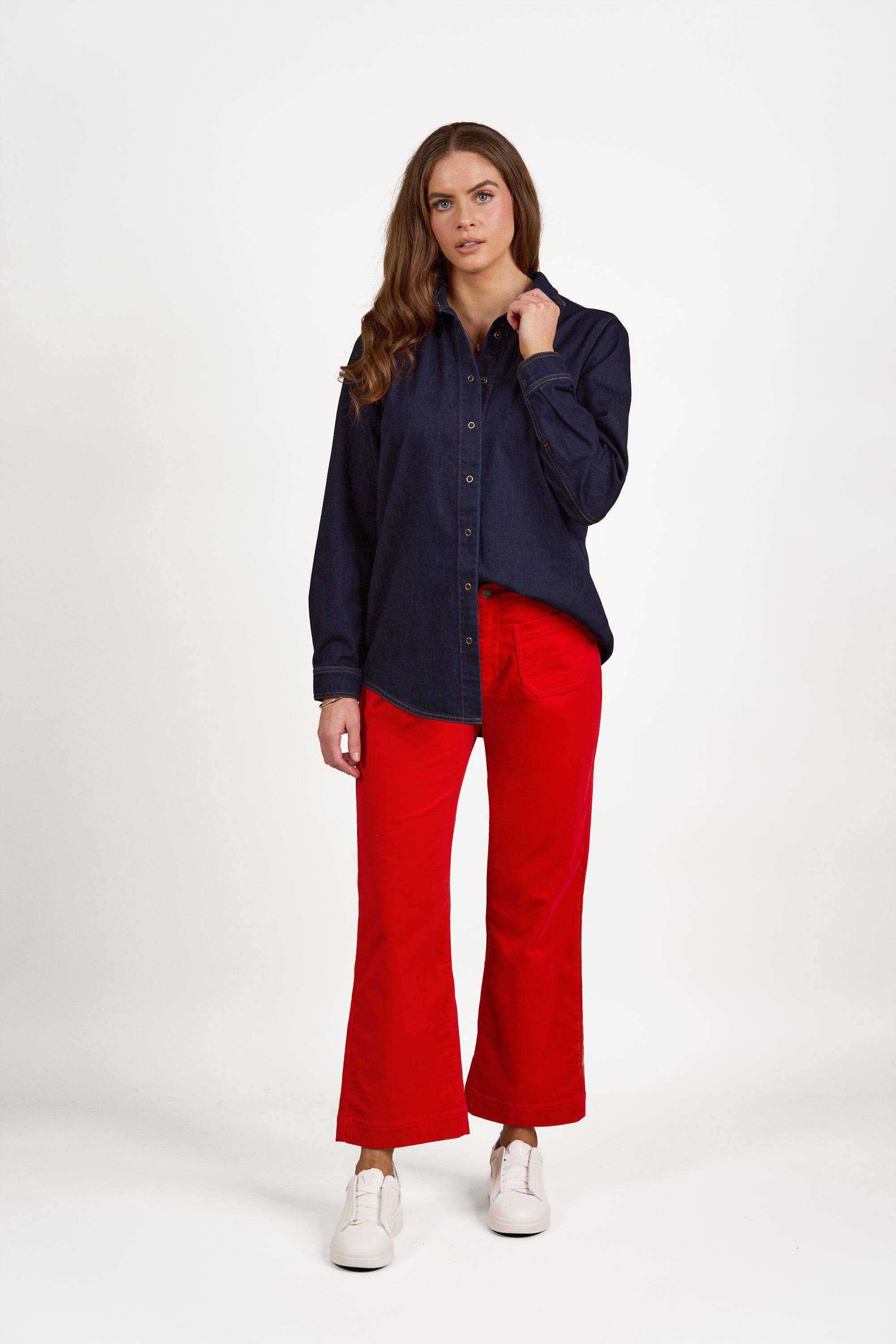 A person stands against a white background wearing the Vassalli Shirt With Poppers in Dark Denim, featuring contrast stitching, paired with bright red pants and white sneakers. They have one hand touching their collar and a neutral expression on their face.