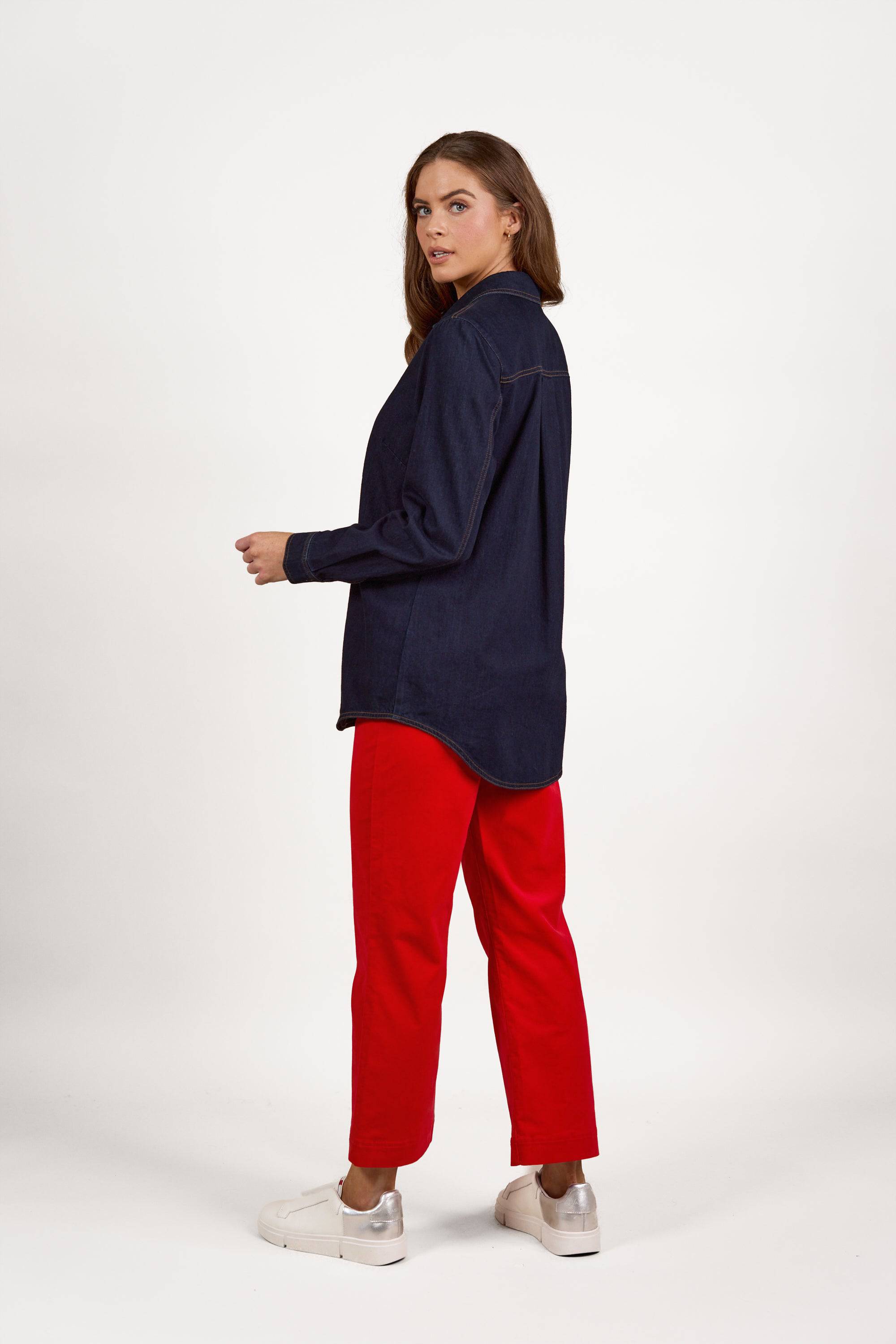 A woman stands sideways, glancing over her shoulder. She is wearing a slim-fit, long-sleeve Shirt With Poppers – Dark Denim by Vassalli, featuring contrast stitching, along with bright red pants and white platform sneakers. The background is plain white.