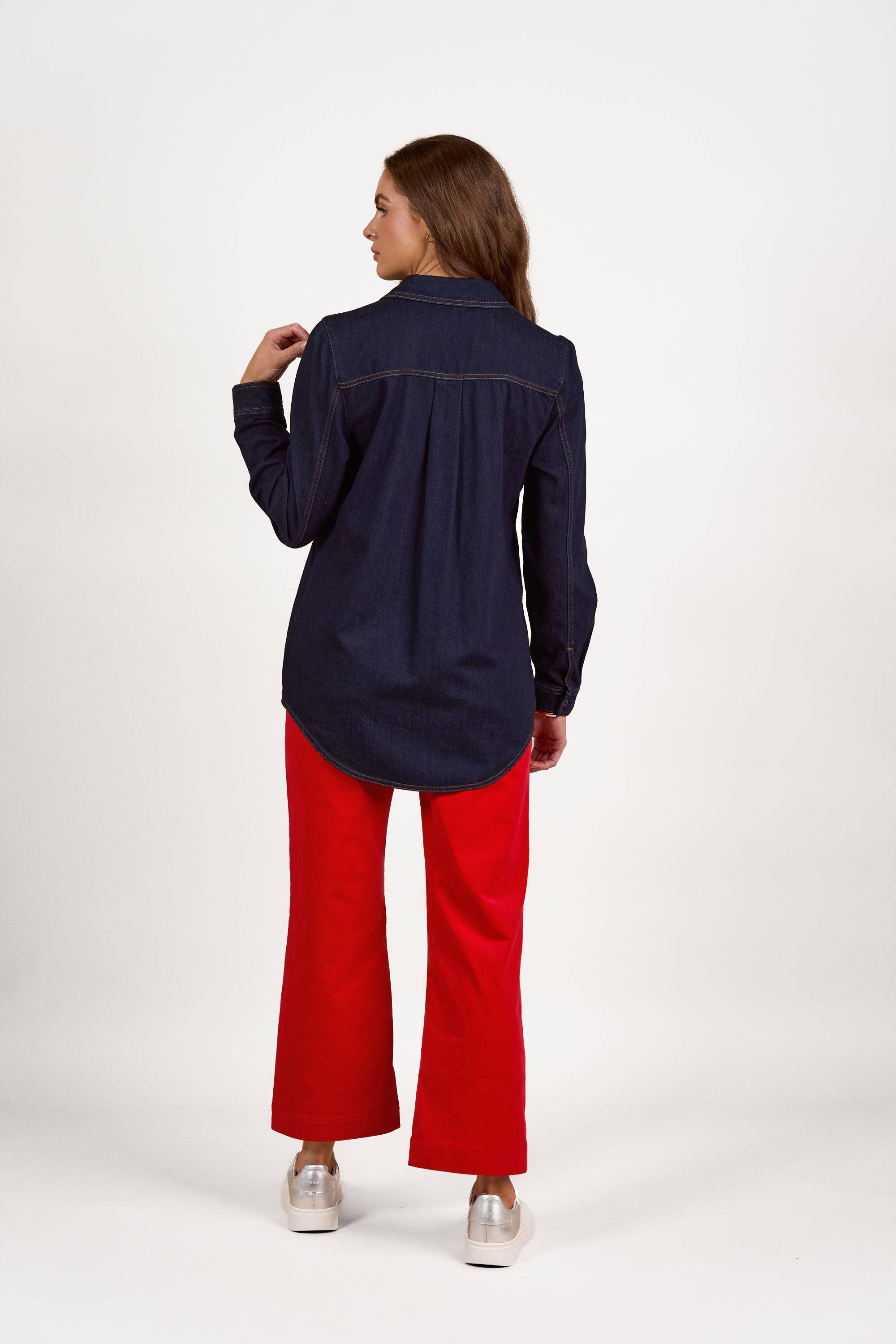 A woman stands facing away, wearing the Vassalli Shirt With Poppers in dark denim with contrast stitching, paired with bright red wide-leg pants and light-colored shoes against a white background.