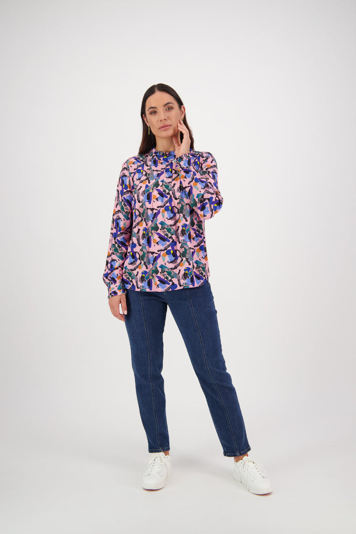 A woman with long dark hair touches her face while wearing the Vassalli Ruffle Neck Top - Trend, blue jeans, and white sneakers against a plain white background.