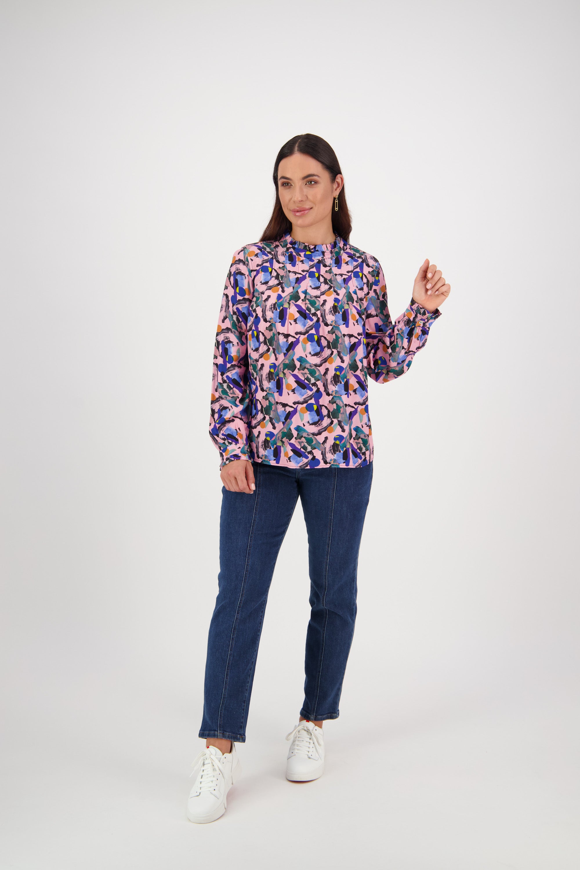 A woman with straight dark hair stands against a white background wearing a Vassalli Ruffle Neck Top - Trend, blue jeans, and white sneakers. She looks slightly to the side with a relaxed expression.
