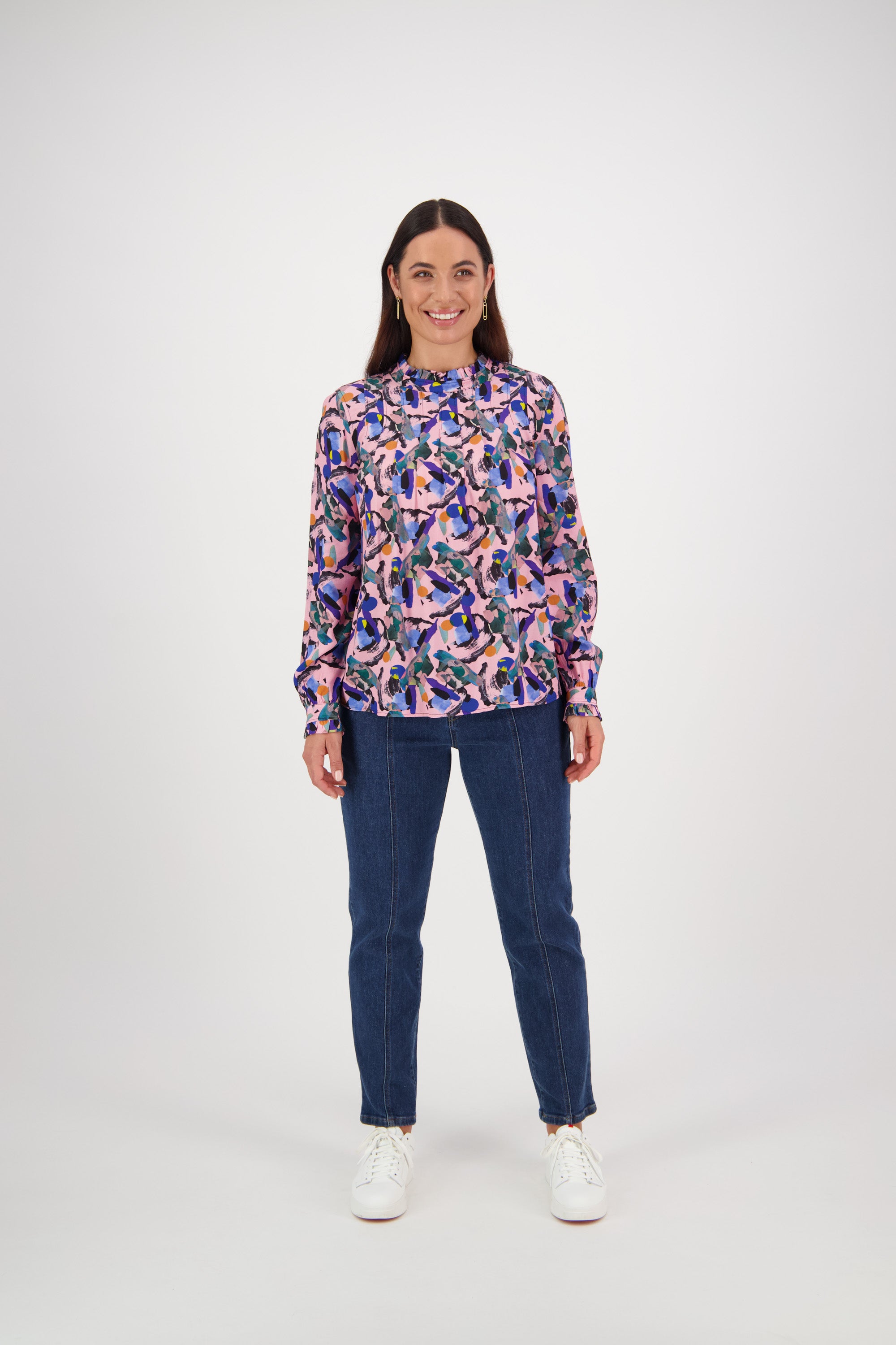 A woman smiles against a plain white background, wearing a Vassalli Ruffle Neck Top - Trend, paired with blue jeans and white sneakers.