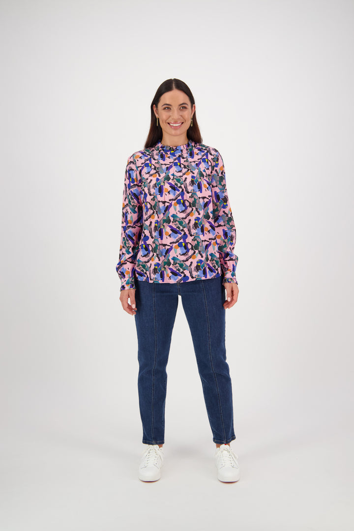 A woman smiles against a plain white background, wearing a Vassalli Ruffle Neck Top - Trend, paired with blue jeans and white sneakers.