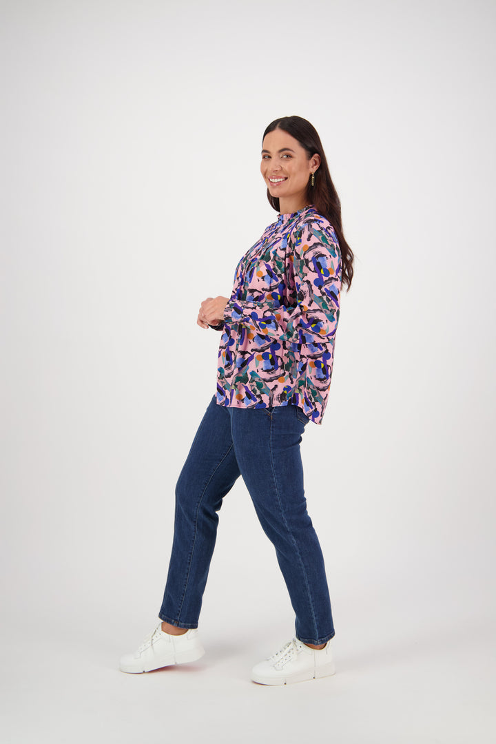 A woman smiles as she poses at an angle against a plain white background, wearing a Vassalli Ruffle Neck Top - Trend, blue jeans, and white sneakers.