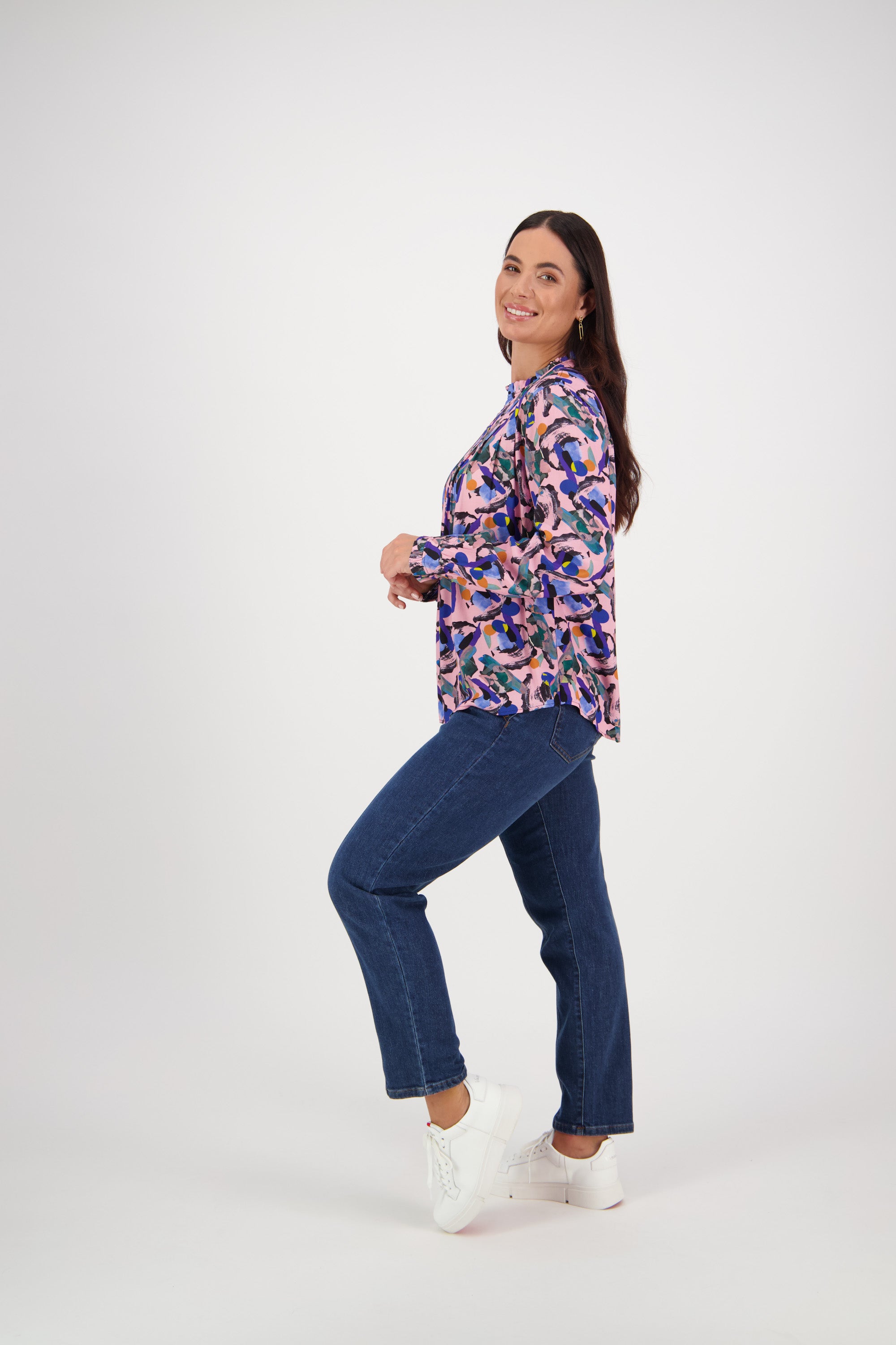 A woman poses side-on, smiling with one leg raised. She wears the Vassalli Ruffle Neck Top - Trend, blue jeans, and white sneakers, set against a plain white background.