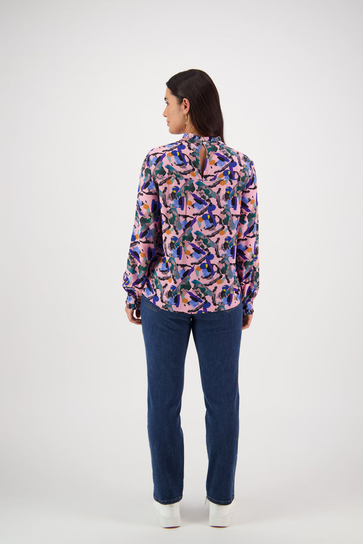 A woman faces away from the camera, wearing a Vassalli Ruffle Neck Top - Trend with blue jeans and white shoes, set against a plain white background.