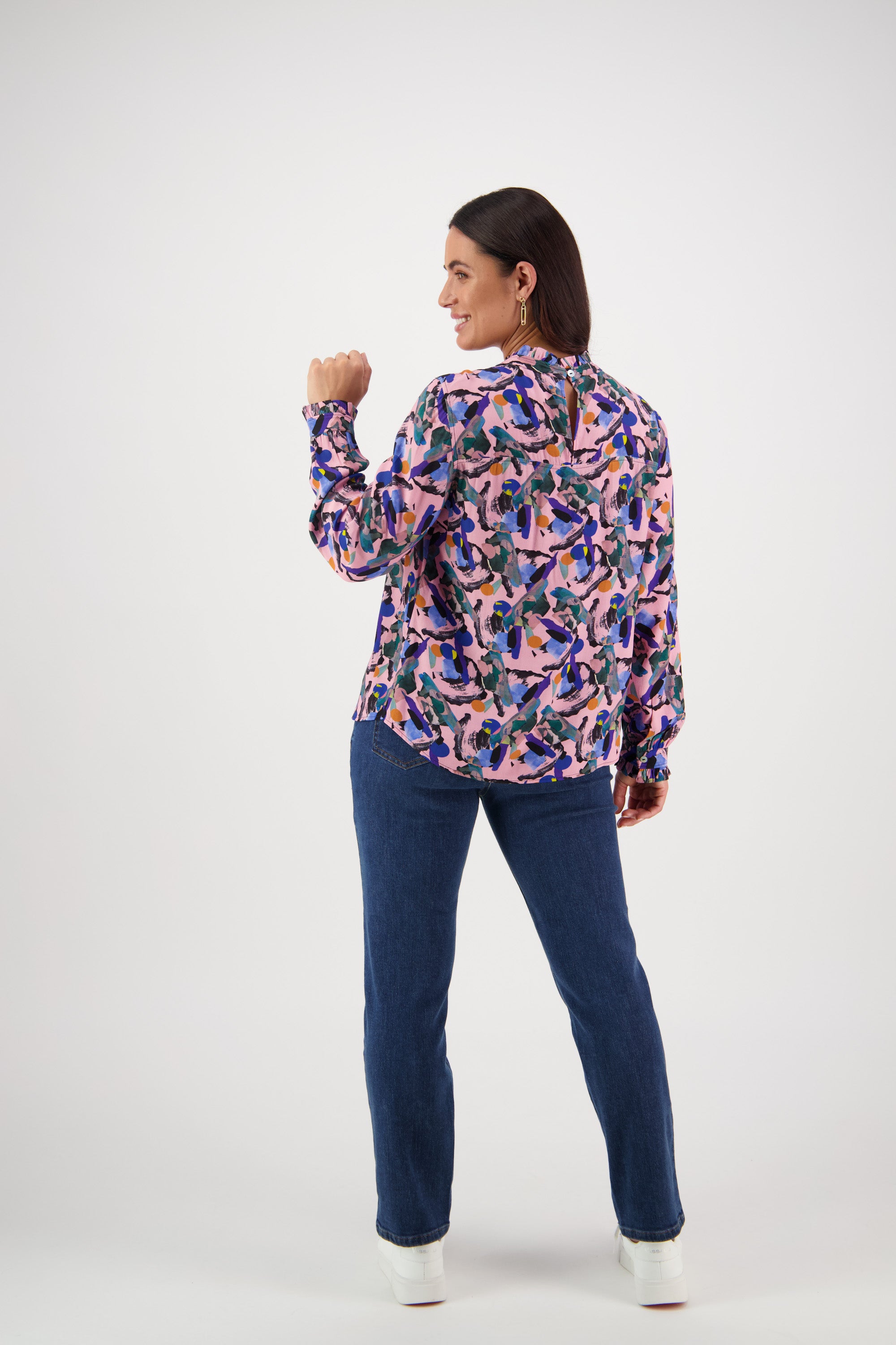 A woman stands facing away, turning to smile. She wears a Vassalli Ruffle Neck Top - Trend with an exclusive abstract print, long sleeves, blue jeans, and white shoes against a plain white background.