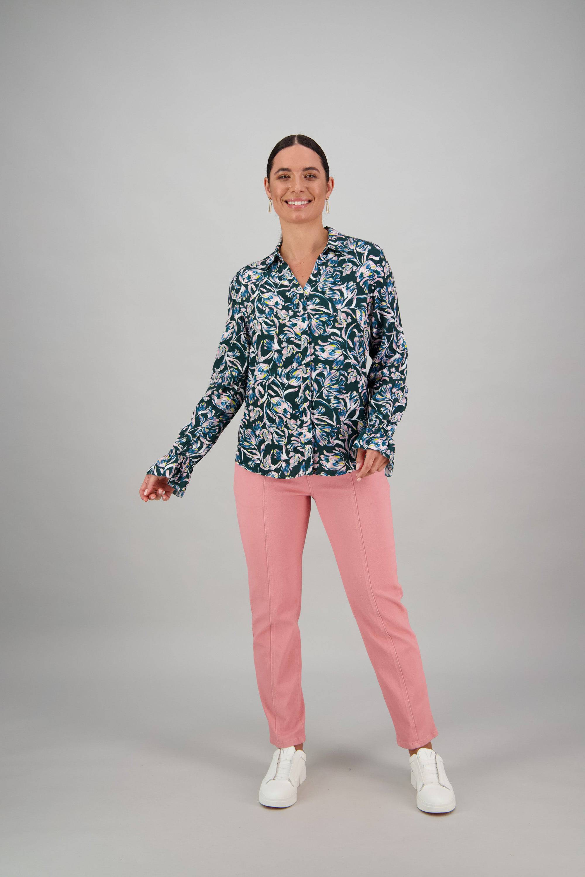 A woman with dark hair pulled back smiles confidently against a plain gray background, modeling the Vassalli Long Sleeve Shirt - Valentina. She pairs it with pink pants and white sneakers, one hand relaxed by her side.