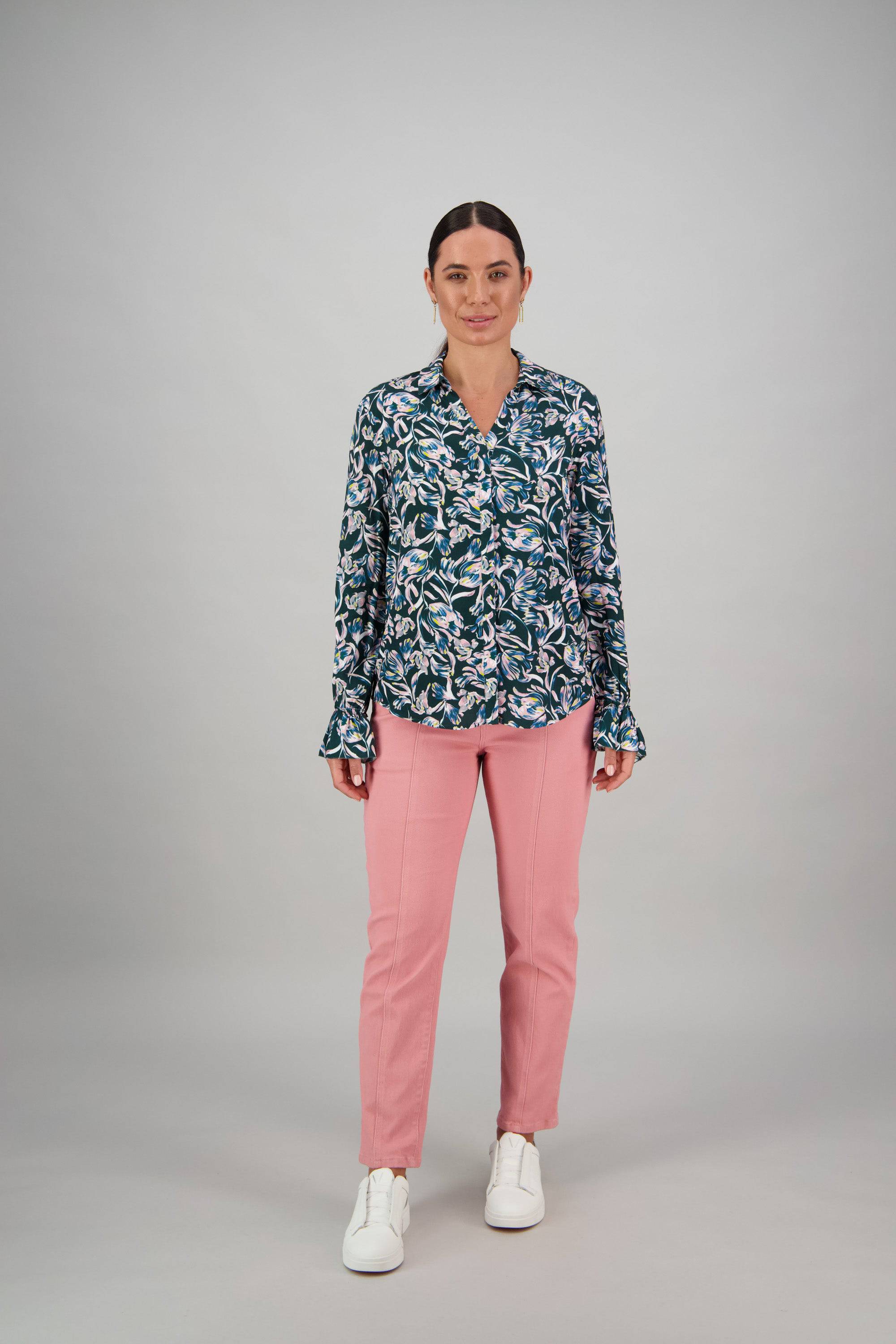 A woman with dark hair tied back stands facing forward against a plain light gray background, wearing a blue and white floral Vassalli Long Sleeve Shirt - Valentina, ruffle trim cuffs, light pink pants, and white sneakers.