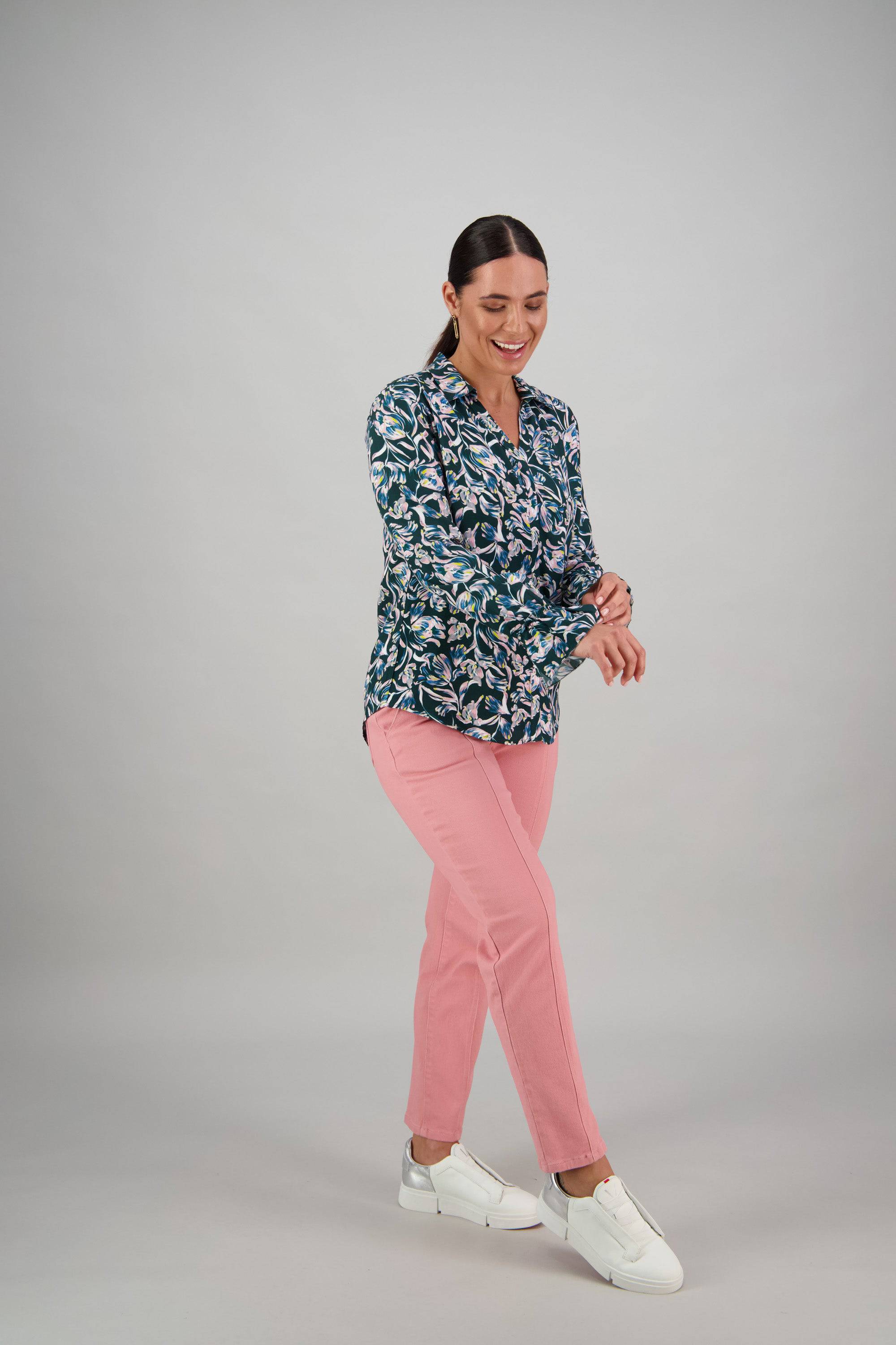 A woman with dark hair in a ponytail wears the Vassalli Long Sleeve Shirt - Valentina with blue florals, pink pants, and white sneakers. She smiles while adjusting her ruffle trim cuff against a plain light gray background.