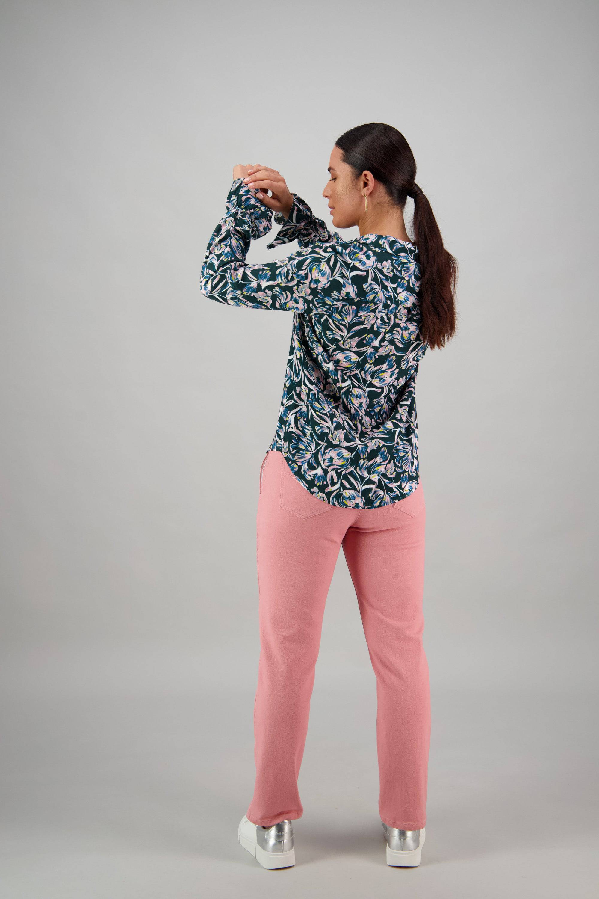 A woman with dark hair in a ponytail, wearing a Vassalli Long Sleeve Shirt - Valentina, pink pants, and white platform shoes stands facing away and adjusts her sleeve against a plain light gray background.