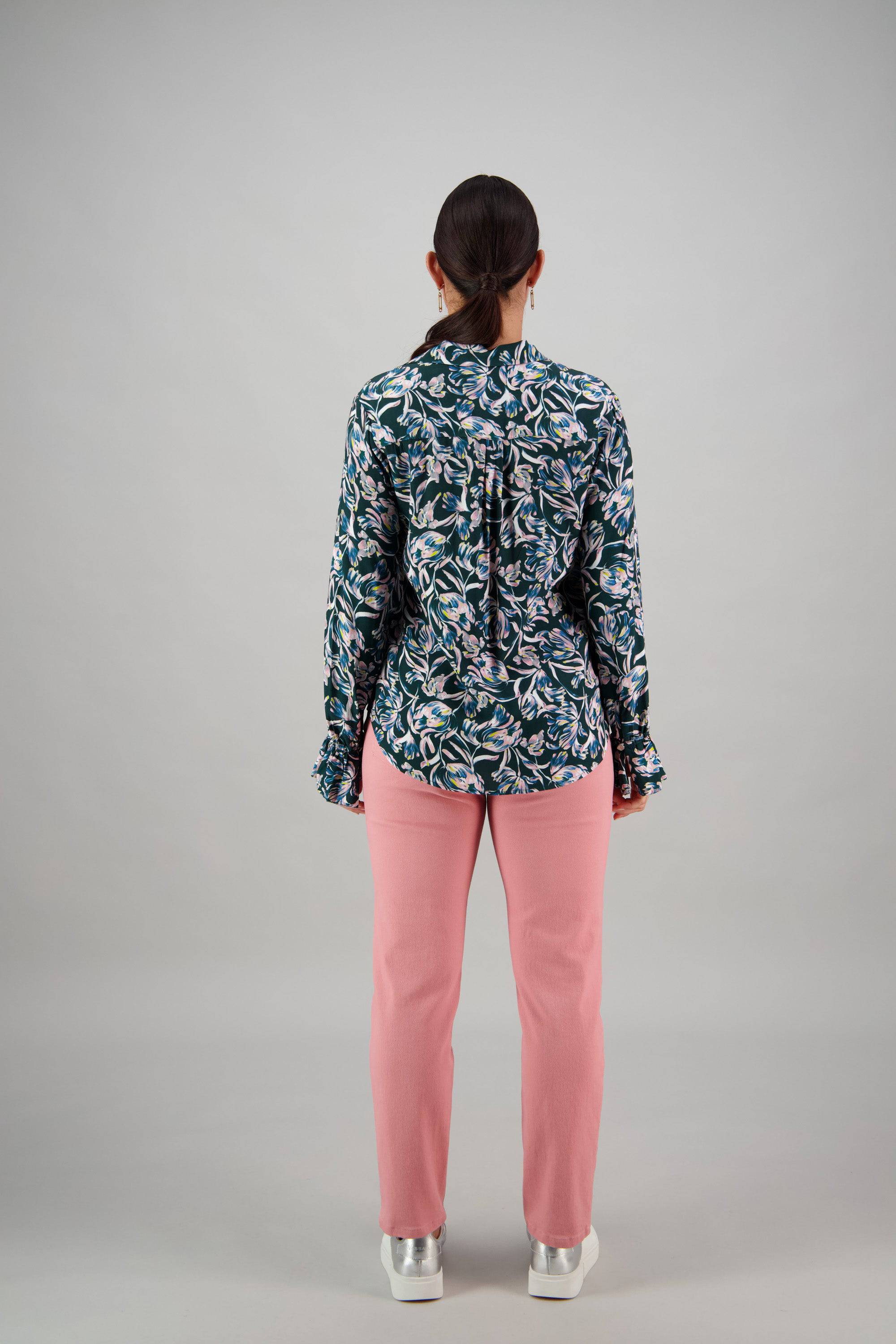 A person with dark hair in a low ponytail stands facing away, wearing the Vassalli Long Sleeve Shirt - Valentina, paired with pink pants and white platform shoes, against a plain light gray background.