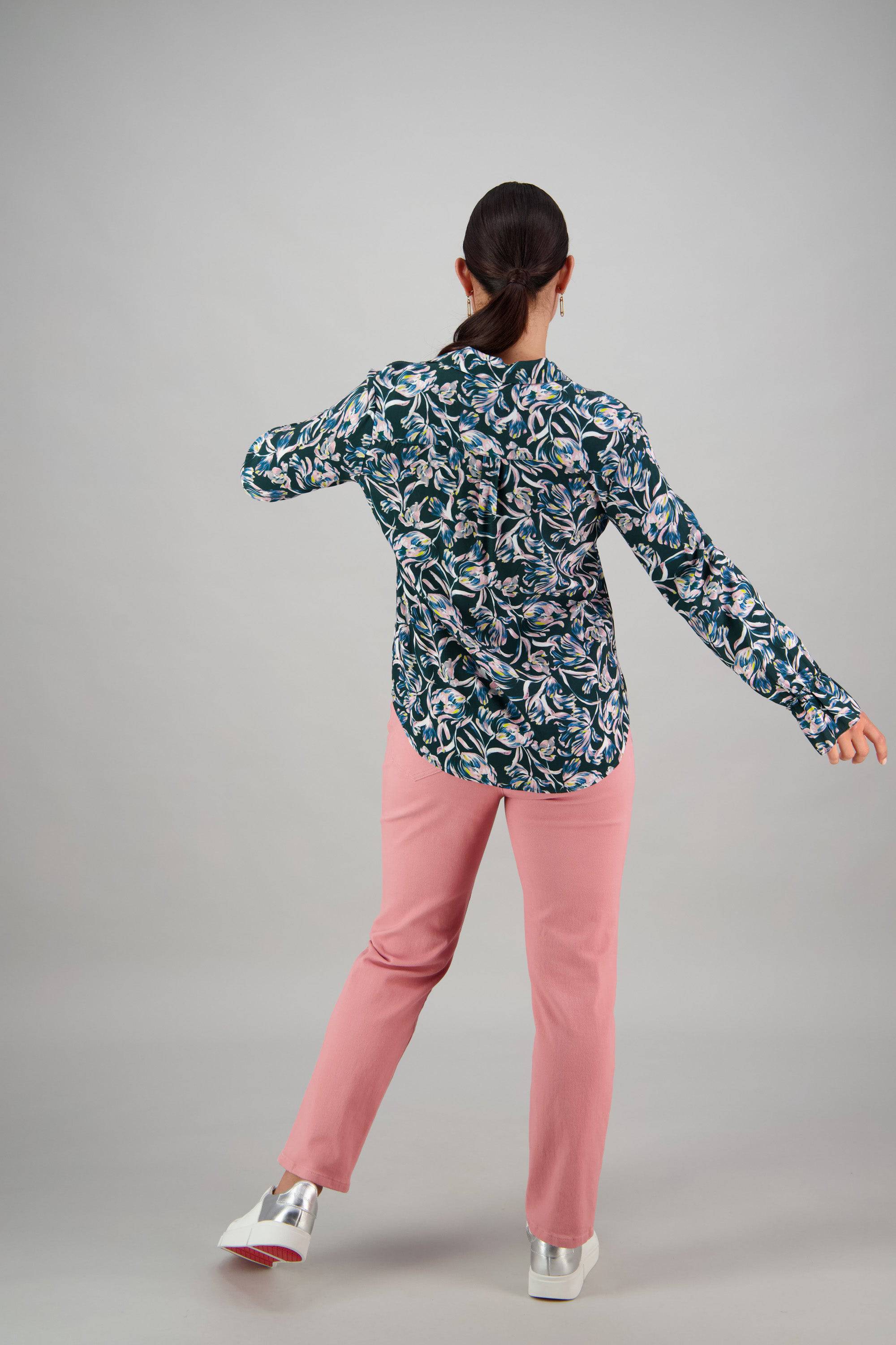 A person with dark hair in a ponytail stands with their back to the camera, wearing a Vassalli Long Sleeve Shirt - Valentina, pink pants, and white shoes, set against a plain gray background.