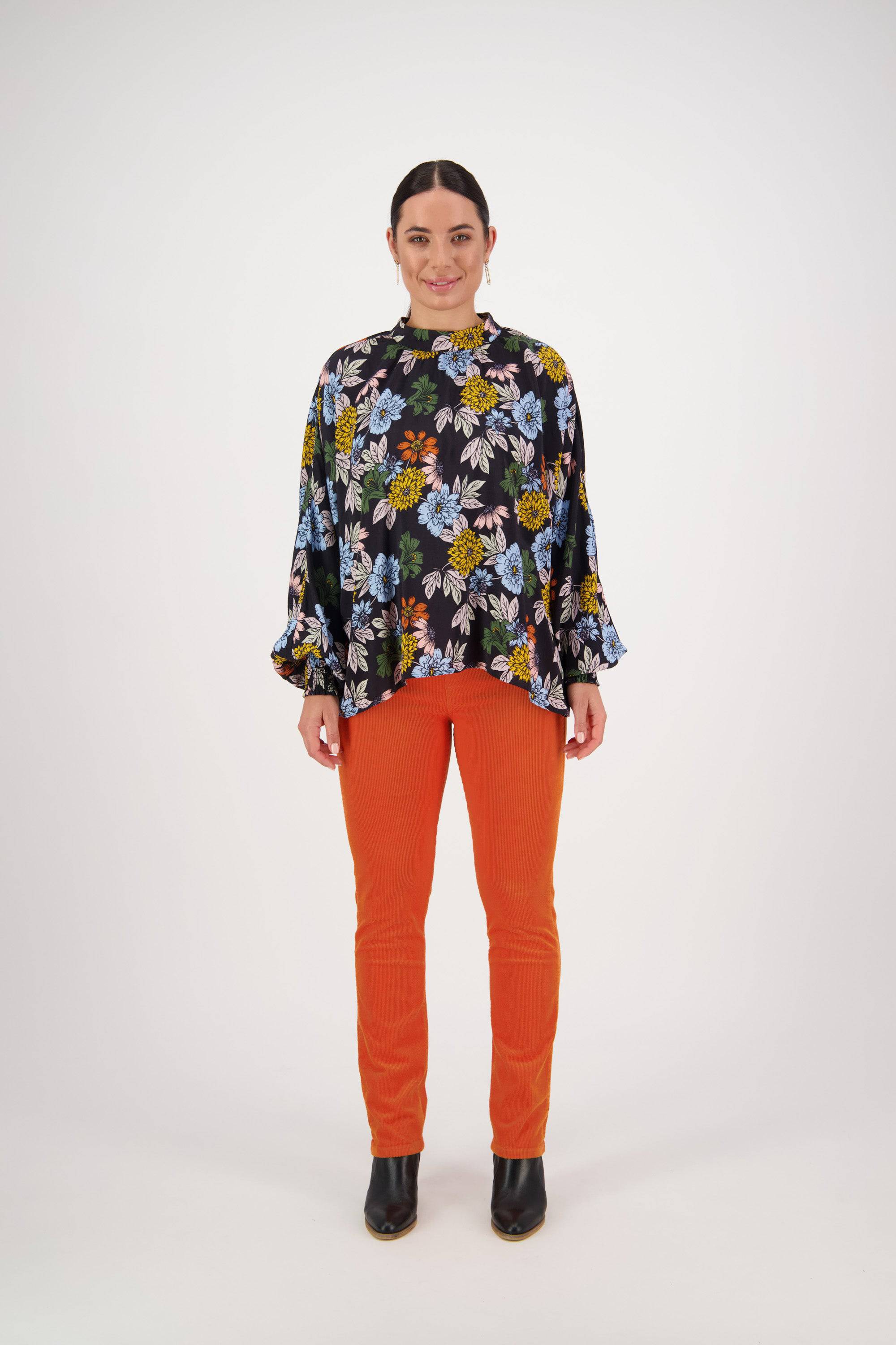 A woman stands facing forward in the Vassalli Printed Top - Viva La Bloom, featuring floaty long sleeves, paired with bright orange pants and black ankle boots, set against a plain white background.