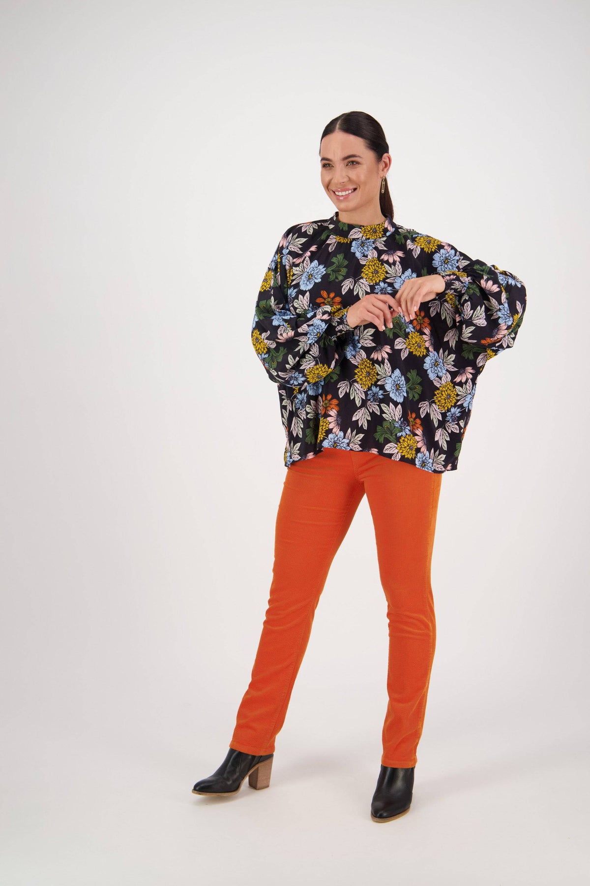 A woman smiles against a plain background in a Vassalli Printed Top - Viva La Bloom, styled with bright orange pants and black heeled ankle boots for a fresh, vibrant look.