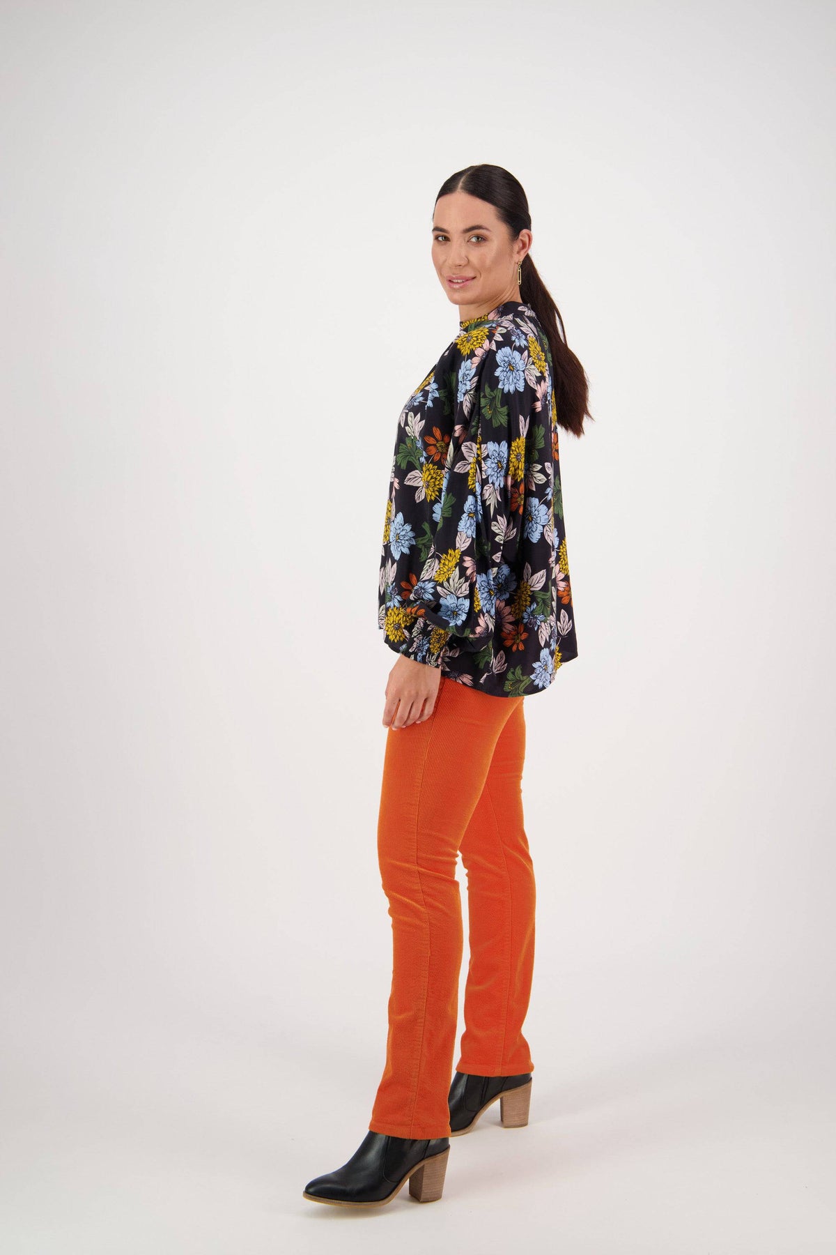A woman stands in a studio wearing the Vassalli Printed Top - Viva La Bloom with floaty long sleeves, bright orange pants, and black heeled boots. She poses smiling with her brown hair in a ponytail against a plain white background.