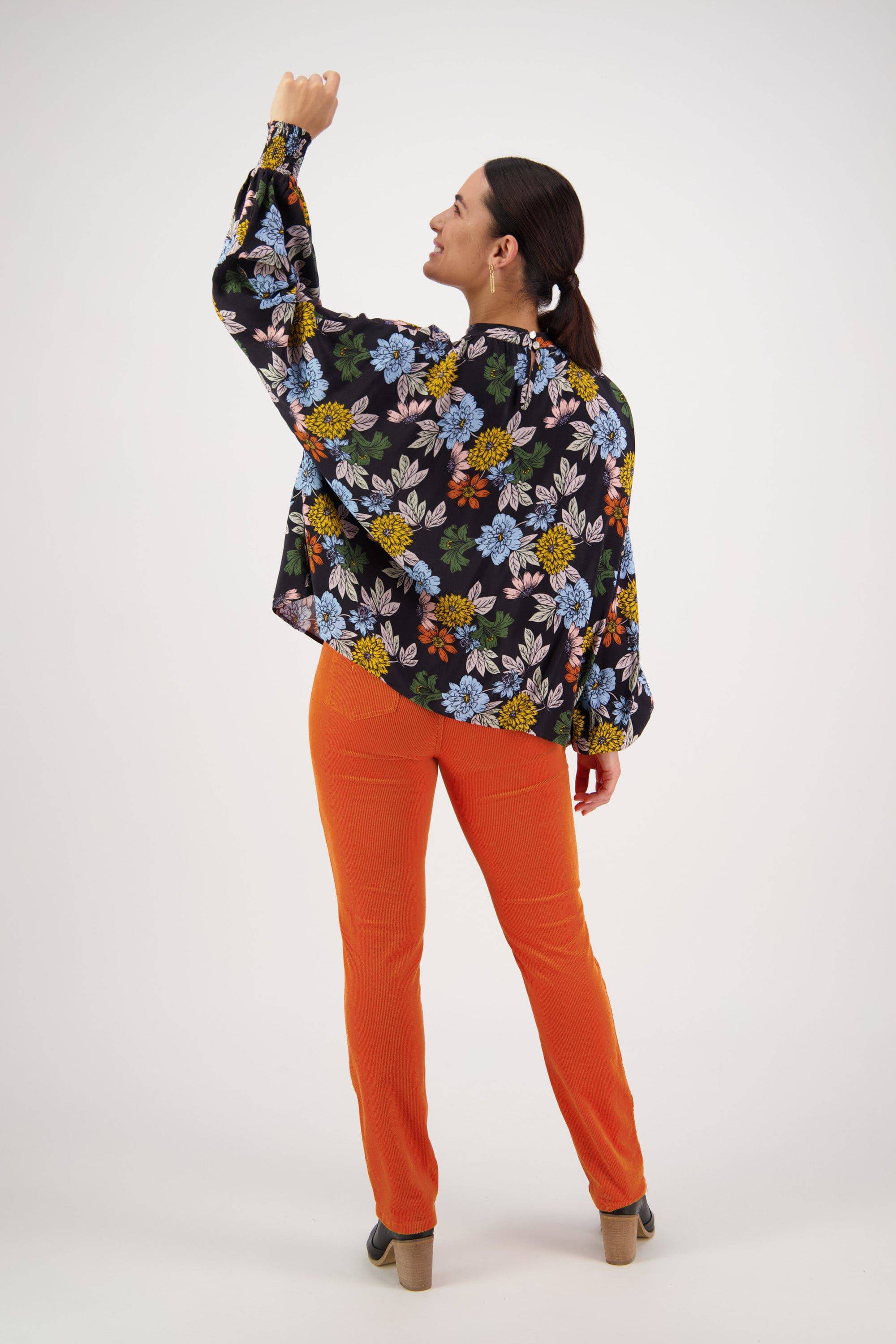 A woman in bright orange pants, heeled boots, and a Vassalli Printed Top - Viva La Bloom with a colorful floral design and floaty long sleeves stands with her back to us, left arm raised, brown hair in a ponytail—radiating Viva La Bloom vibes.