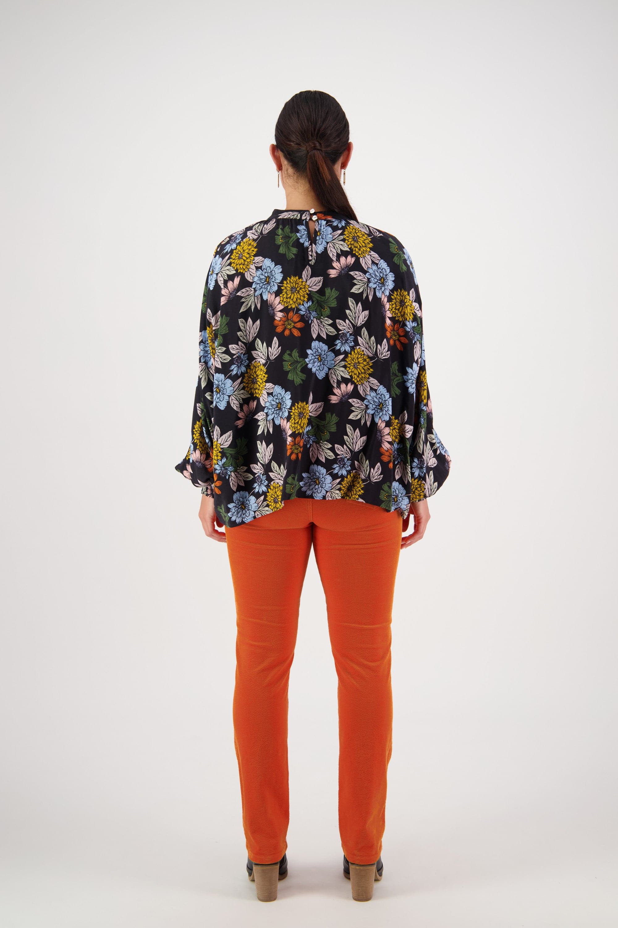 A person faces away from the camera in a Vassalli Printed Top - Viva La Bloom with floaty long sleeves and bright orange pants, set against a plain white background.