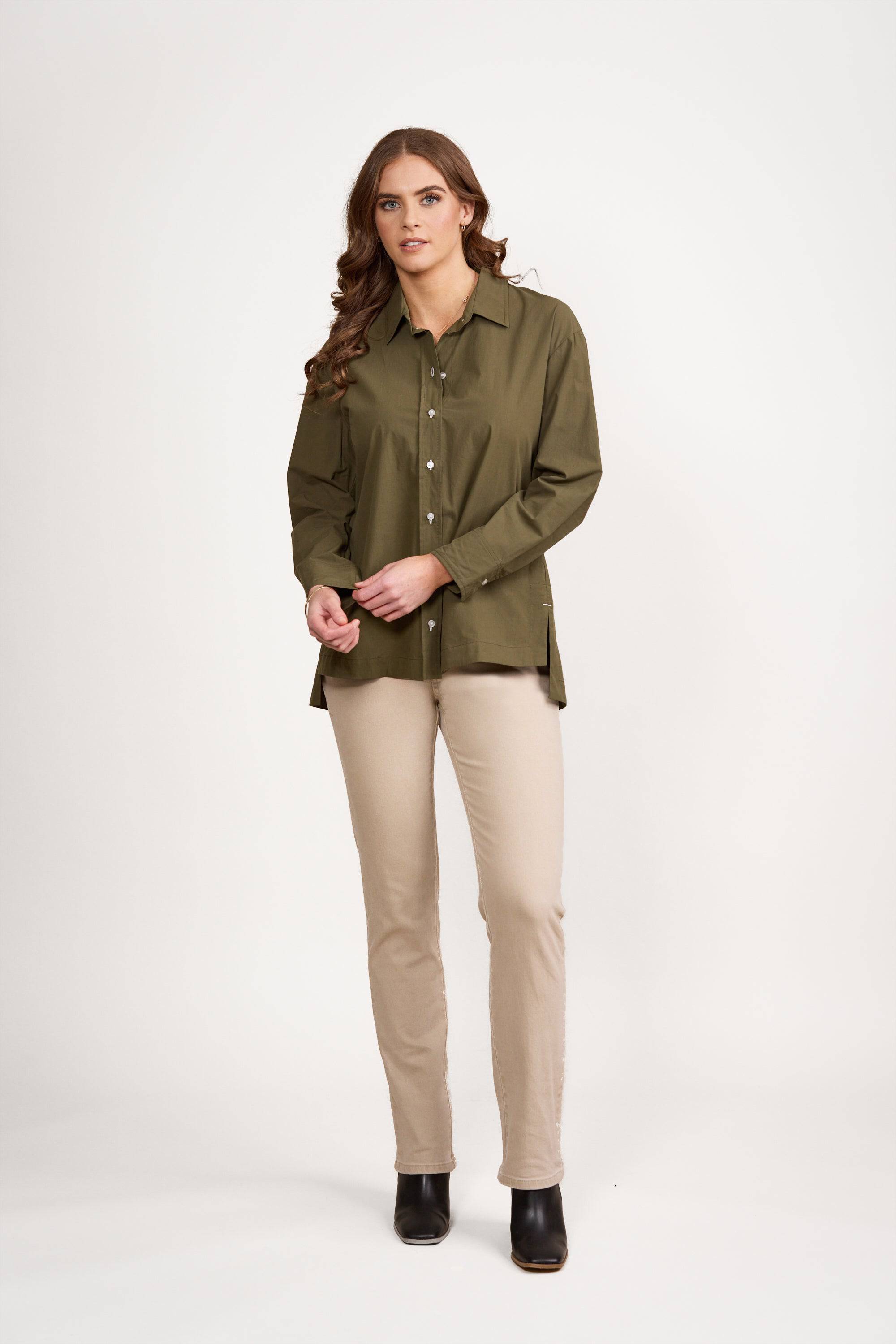 A woman with long brown hair wears the Vassalli Shirt - Olive/White, featuring contrast stitching, paired with beige pants and black ankle boots, standing against a plain white background.