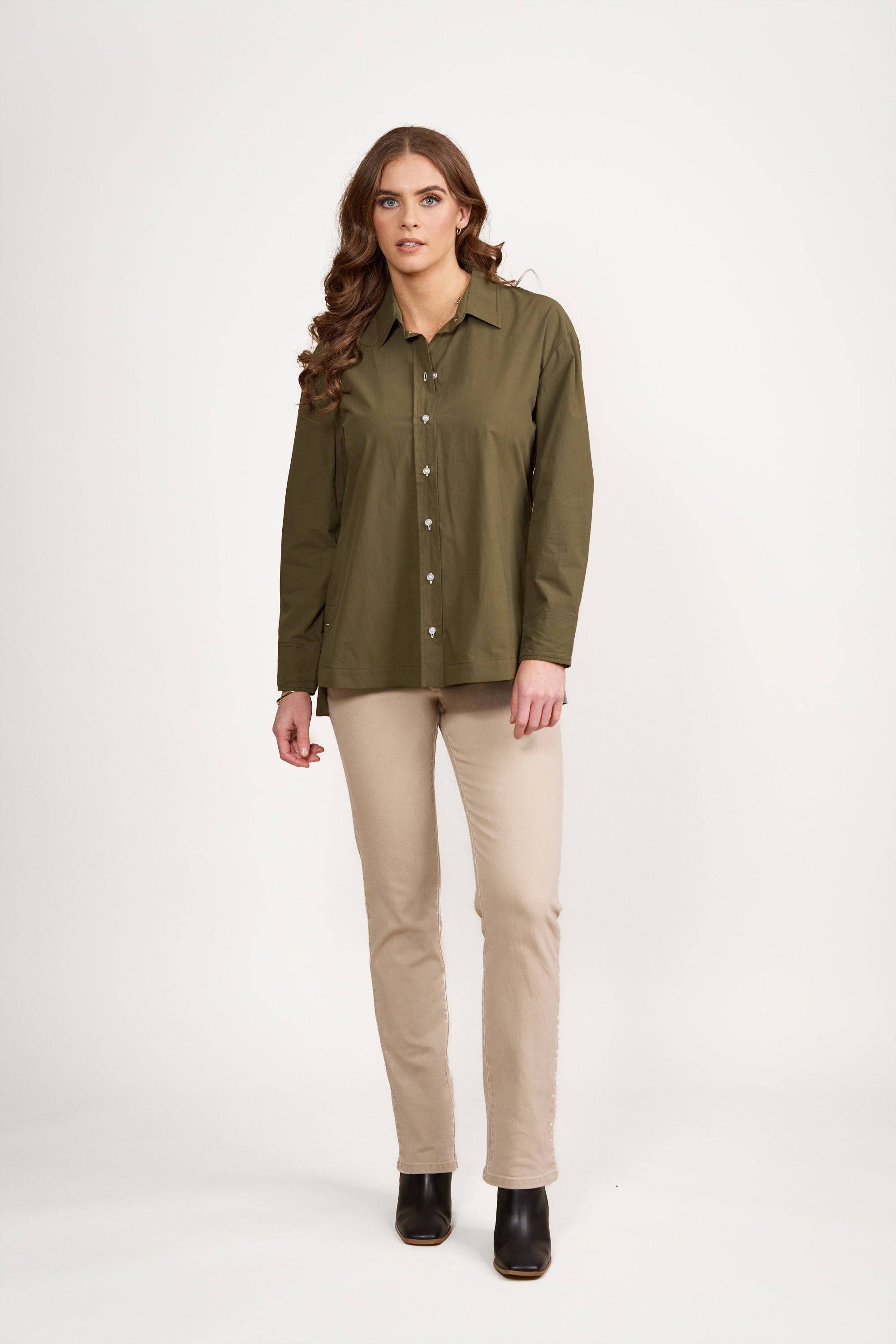 Woman with long brown hair wearing a Vassalli Shirt - Olive/White with contrast stitching, beige pants, and black ankle boots, standing against a plain white background.
