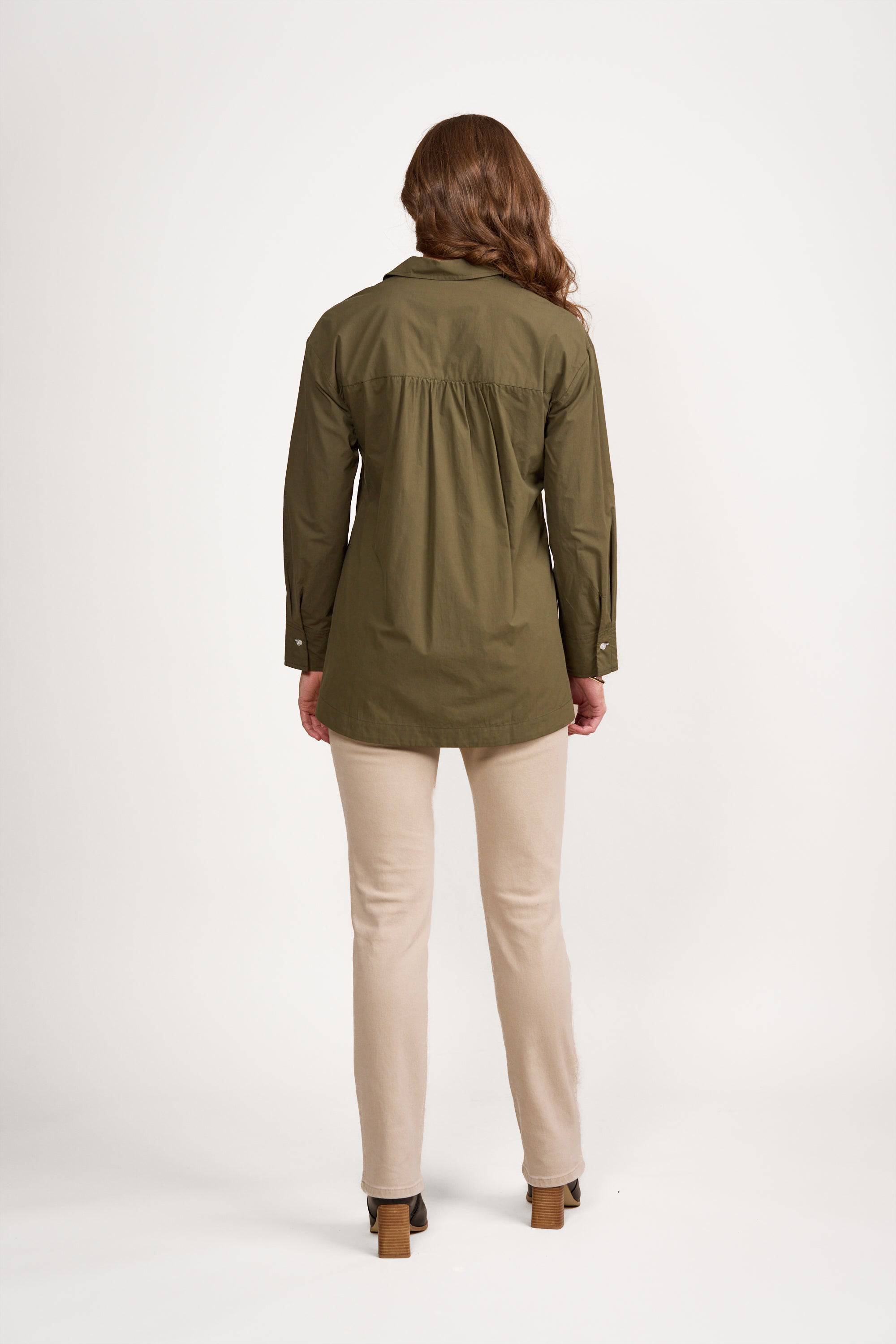 A person with long brown hair stands facing away, wearing the Vassalli Shirt - Olive/White, which features contrast stitching and a loose fit, paired with beige pants and brown heeled shoes, against a plain white background.