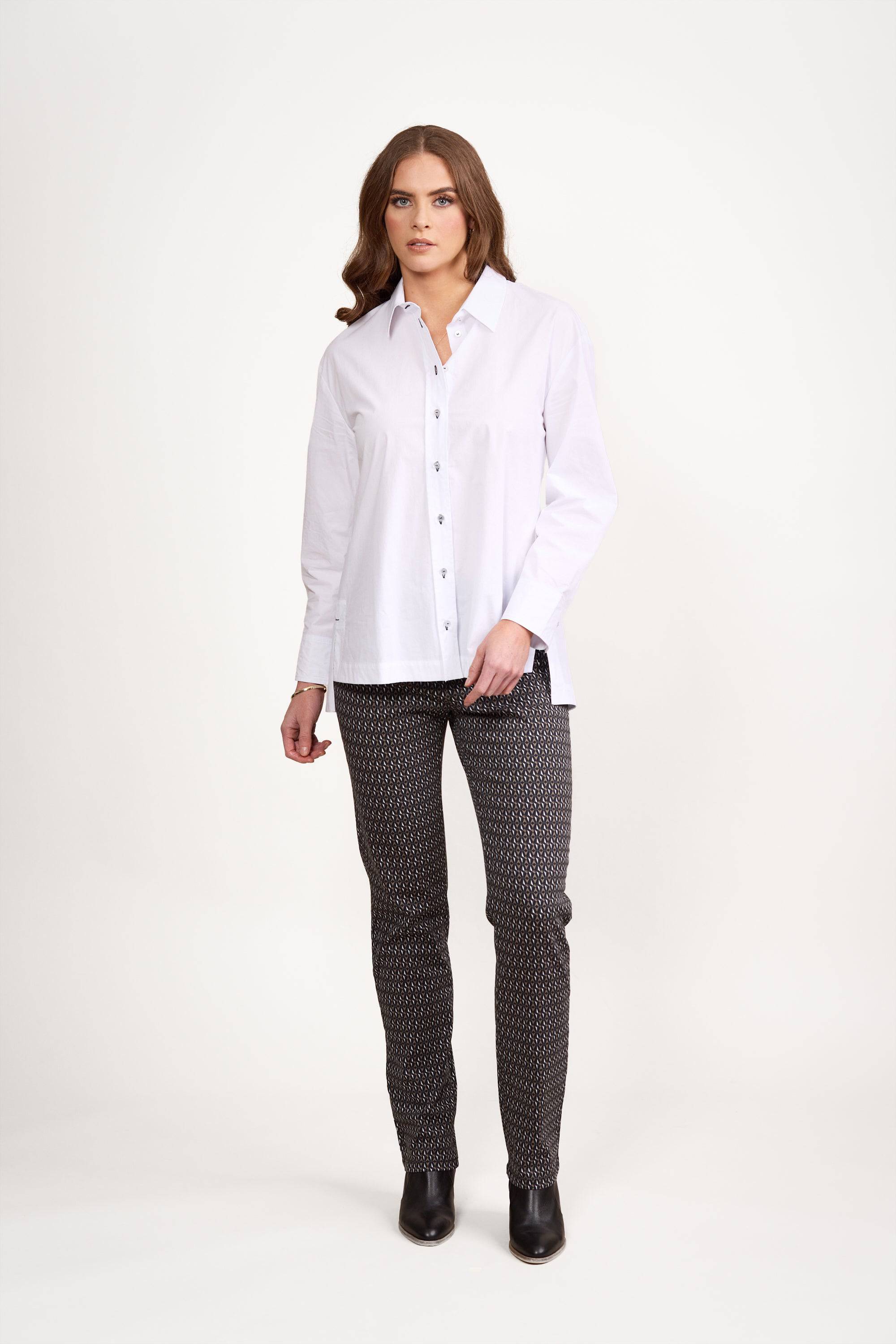 A person stands against a plain background, wearing a loose fit Vassalli Shirt in White/Ink, paired with patterned black pants featuring contrast stitching and black ankle boots.