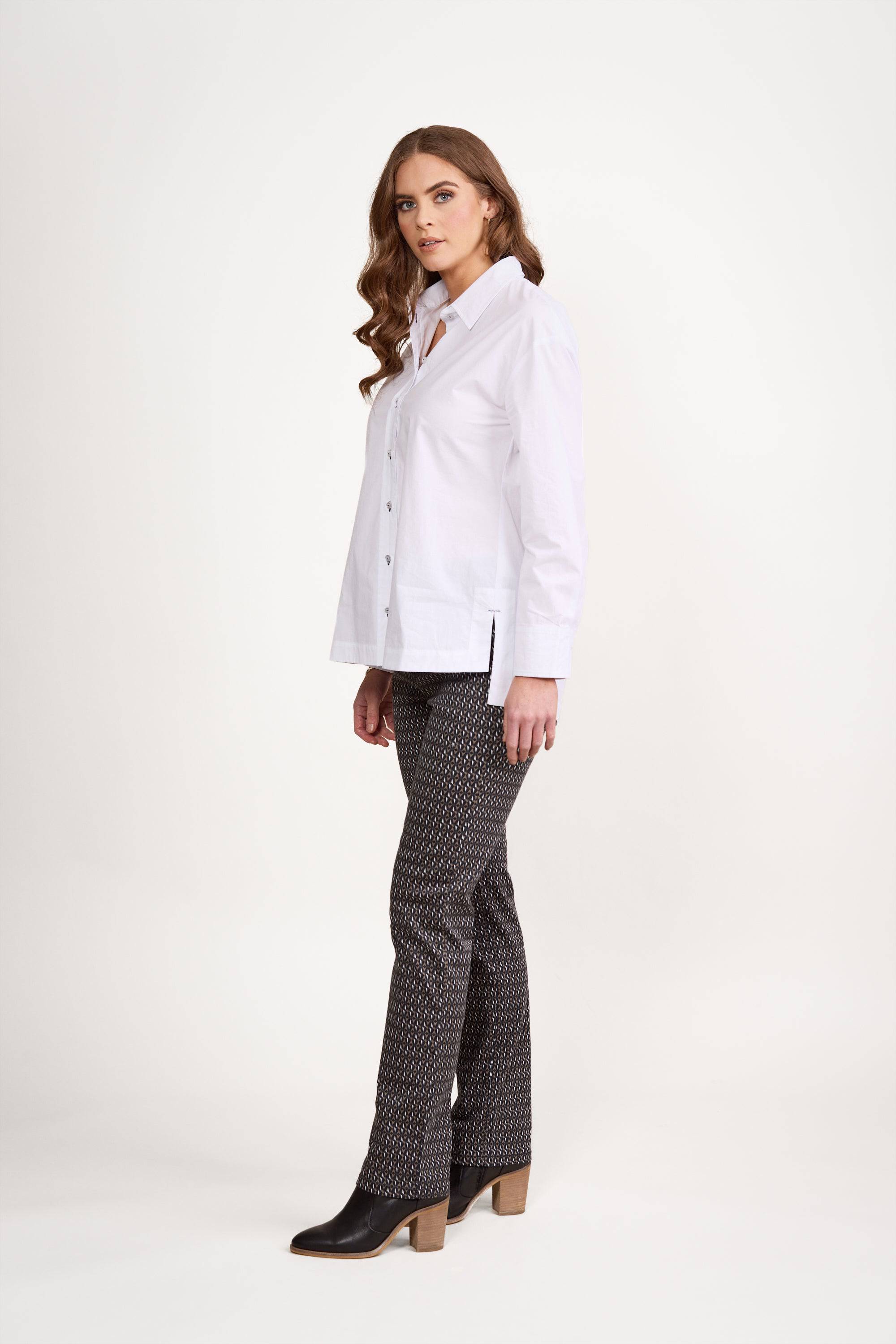 A woman with long wavy hair wears a loose-fit Vassalli Shirt - White/Ink, patterned pants, and black heeled ankle boots, standing sideways against a plain white background.