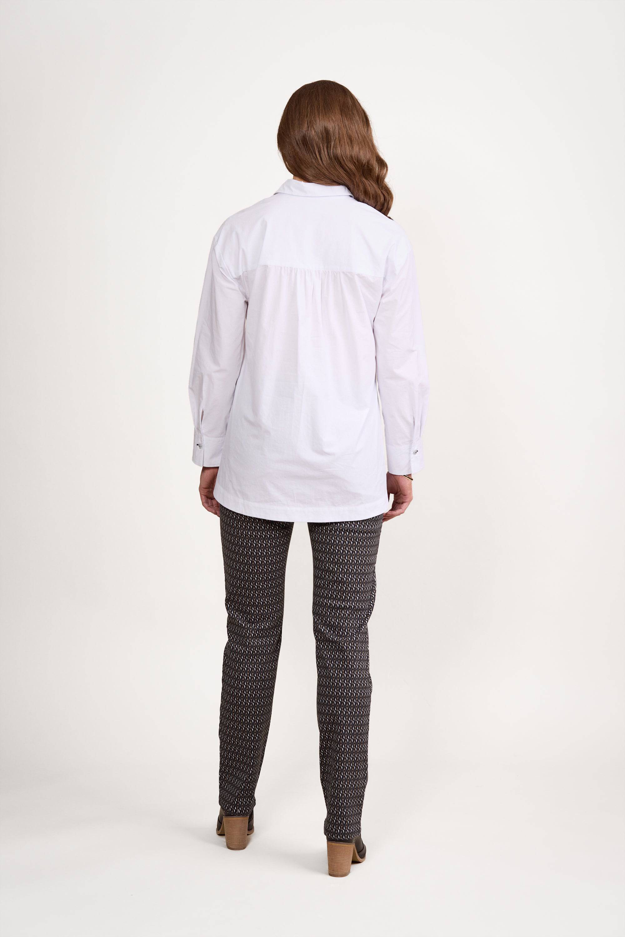 A person with long brown hair stands facing away, wearing a loose-fit Vassalli Shirt - White/Ink and patterned pants with contrast stitching, on a plain white background.