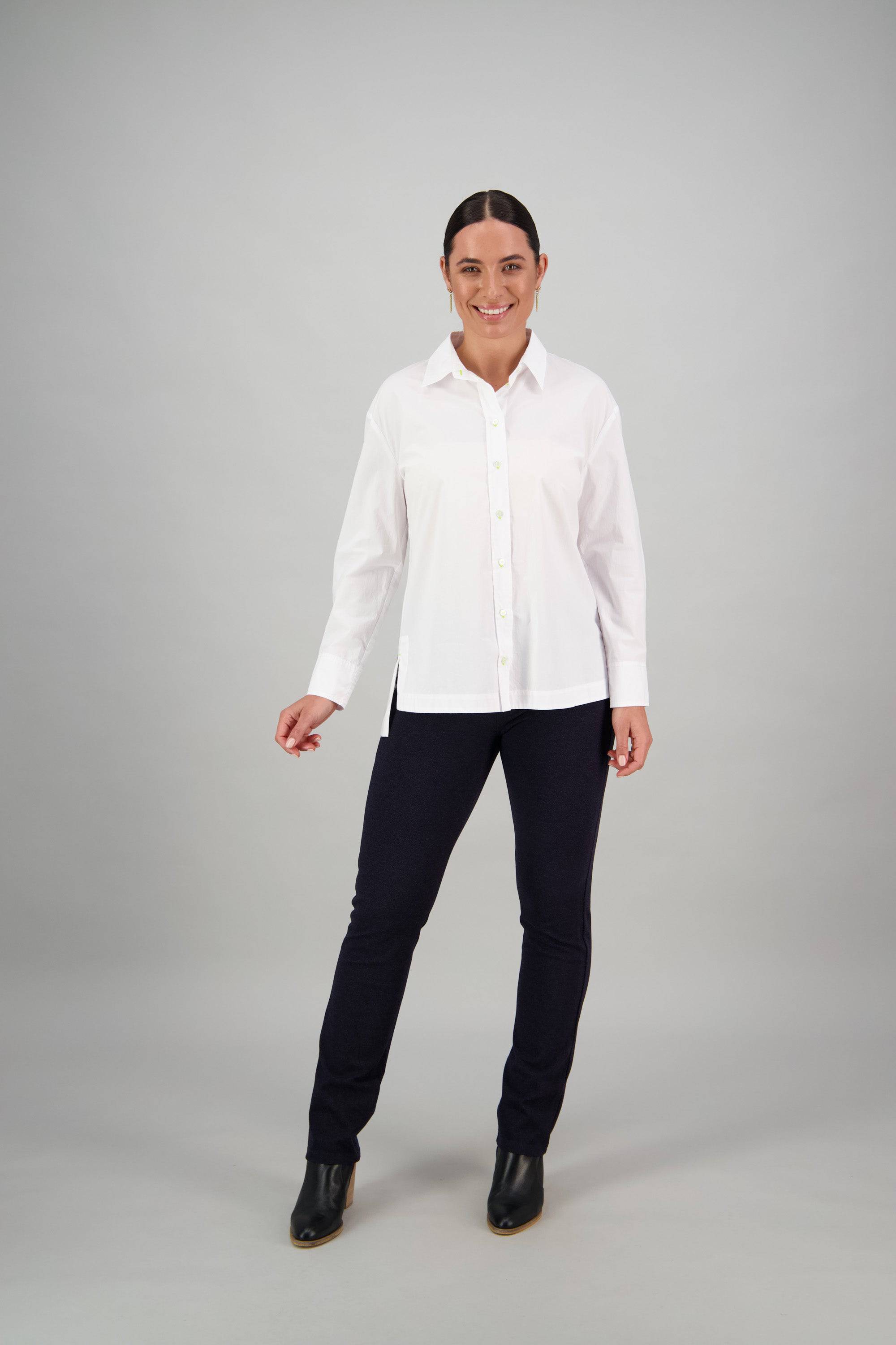 A woman stands smiling, wearing the Vassalli Shirt - White/Lime, paired with black pants and black boots against a plain gray background.