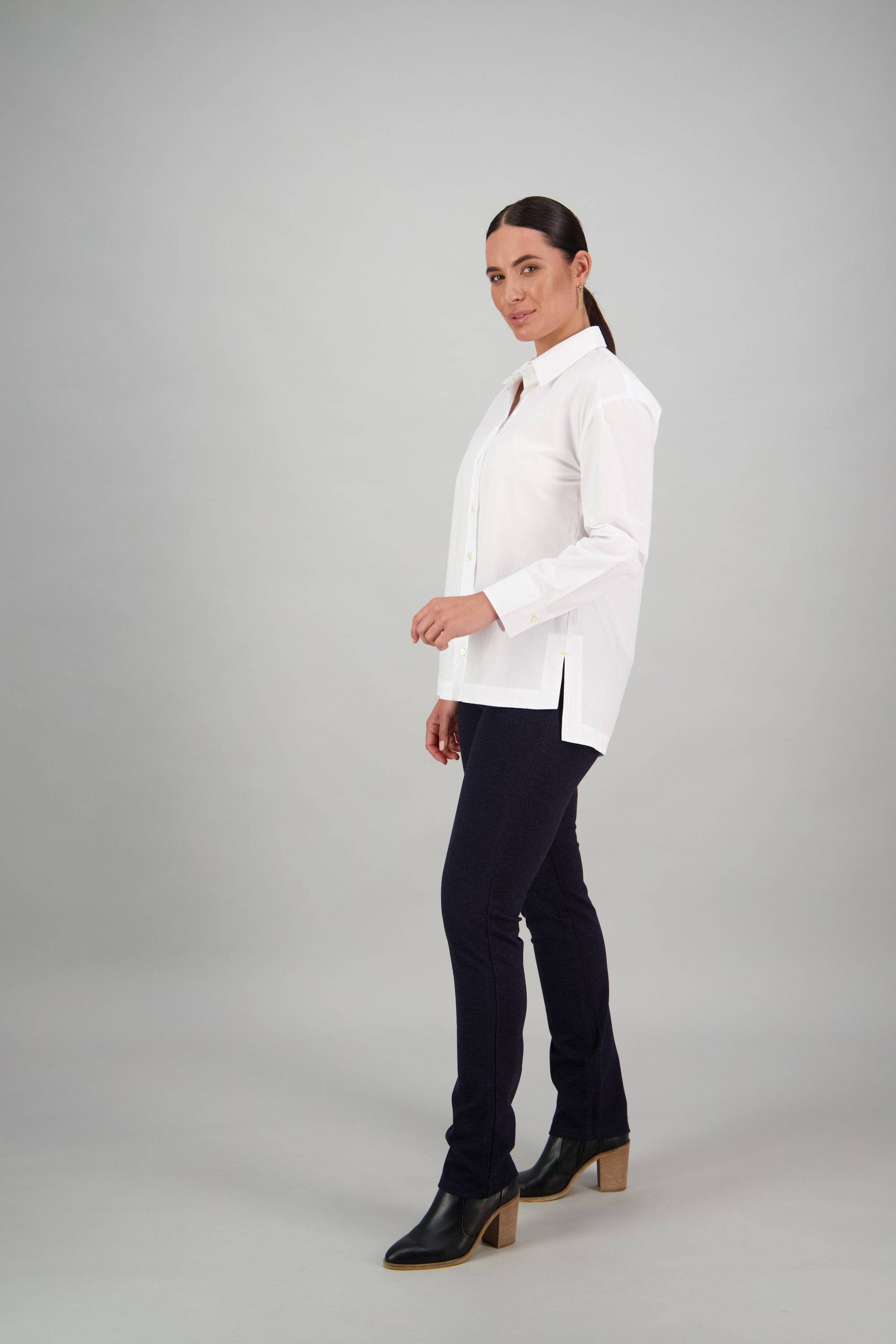 A person stands in a studio wearing the Vassalli Shirt - White/Lime, paired with dark pants and black heeled boots, looking towards the camera with a neutral expression against a plain gray background.
