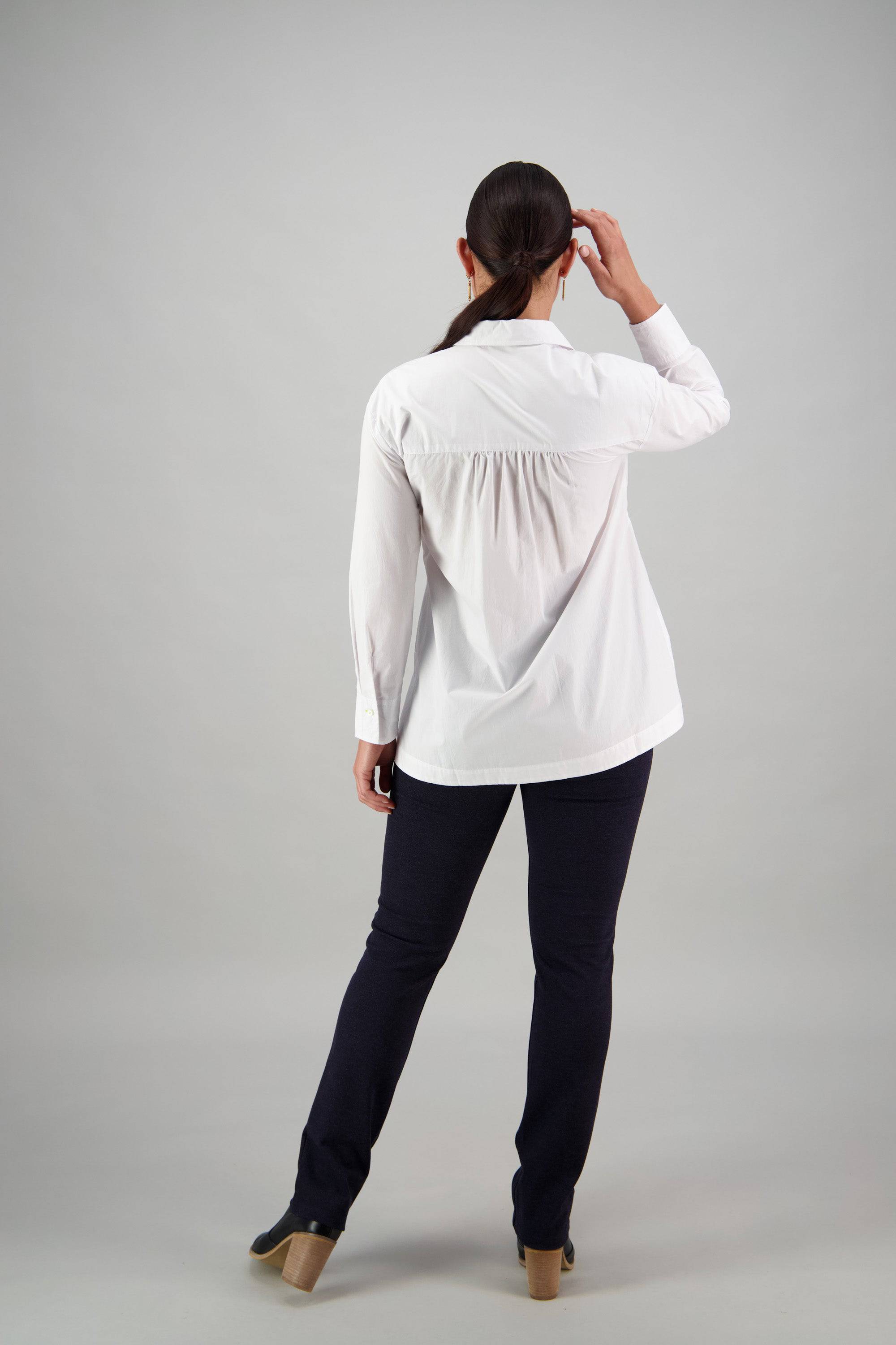 A person with long dark hair, dressed in the Vassalli Shirt - White/Lime and black pants, stands facing away while touching their head with one hand against a plain light gray background.