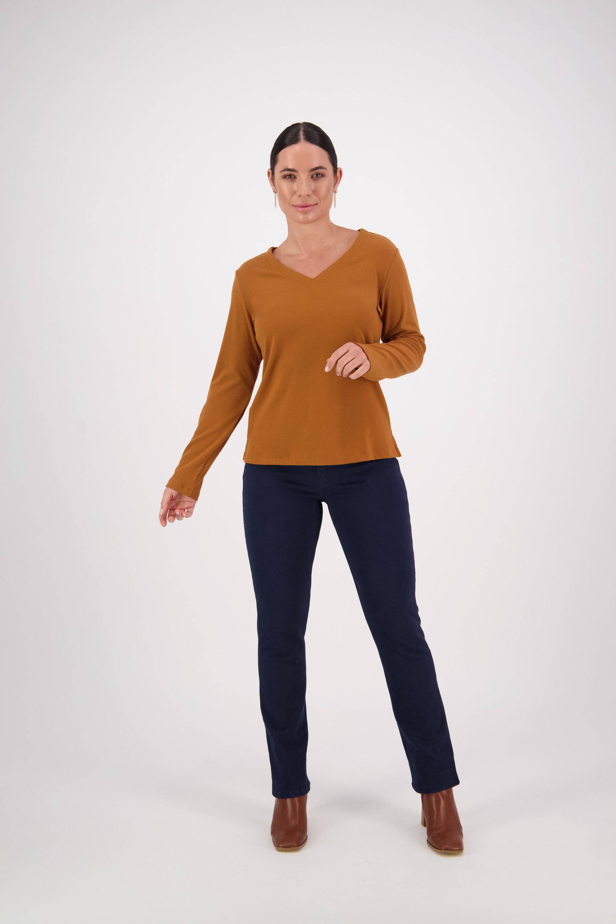 A woman stands against a white background, wearing the Vassalli V-Neck Top in Tobacco, featuring rib fabric and a relaxed fit. She pairs it with dark blue pants and brown ankle boots. Her dark hair is pulled back as she looks forward with a neutral expression, highlighting the comfortable style of her outfit.