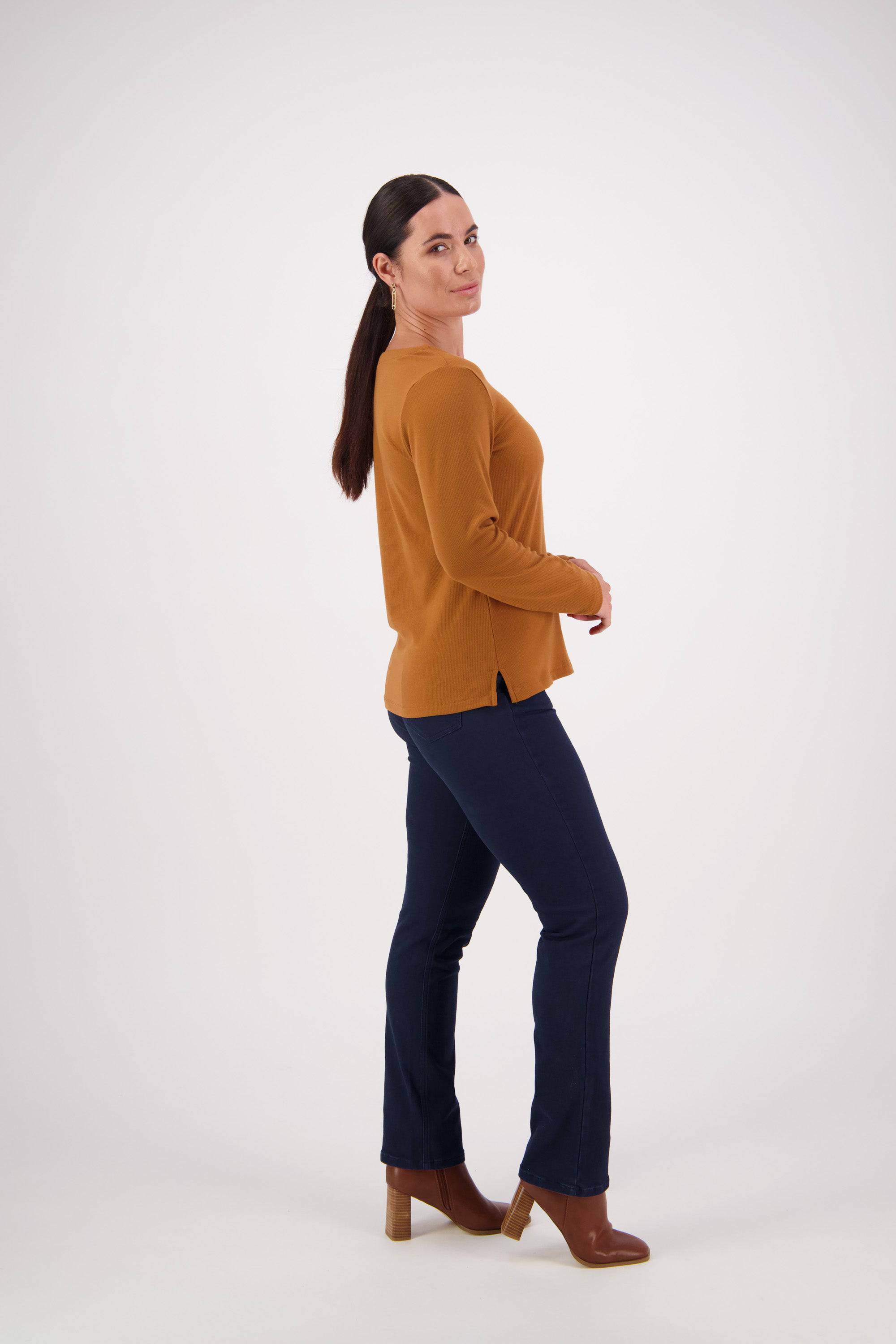 A woman with long dark hair in a ponytail wears a relaxed fit Vassalli V-Neck Top in Tobacco, crafted from rib fabric, paired with dark jeans and brown heeled boots. She stands sideways and smiles slightly against a plain white background.