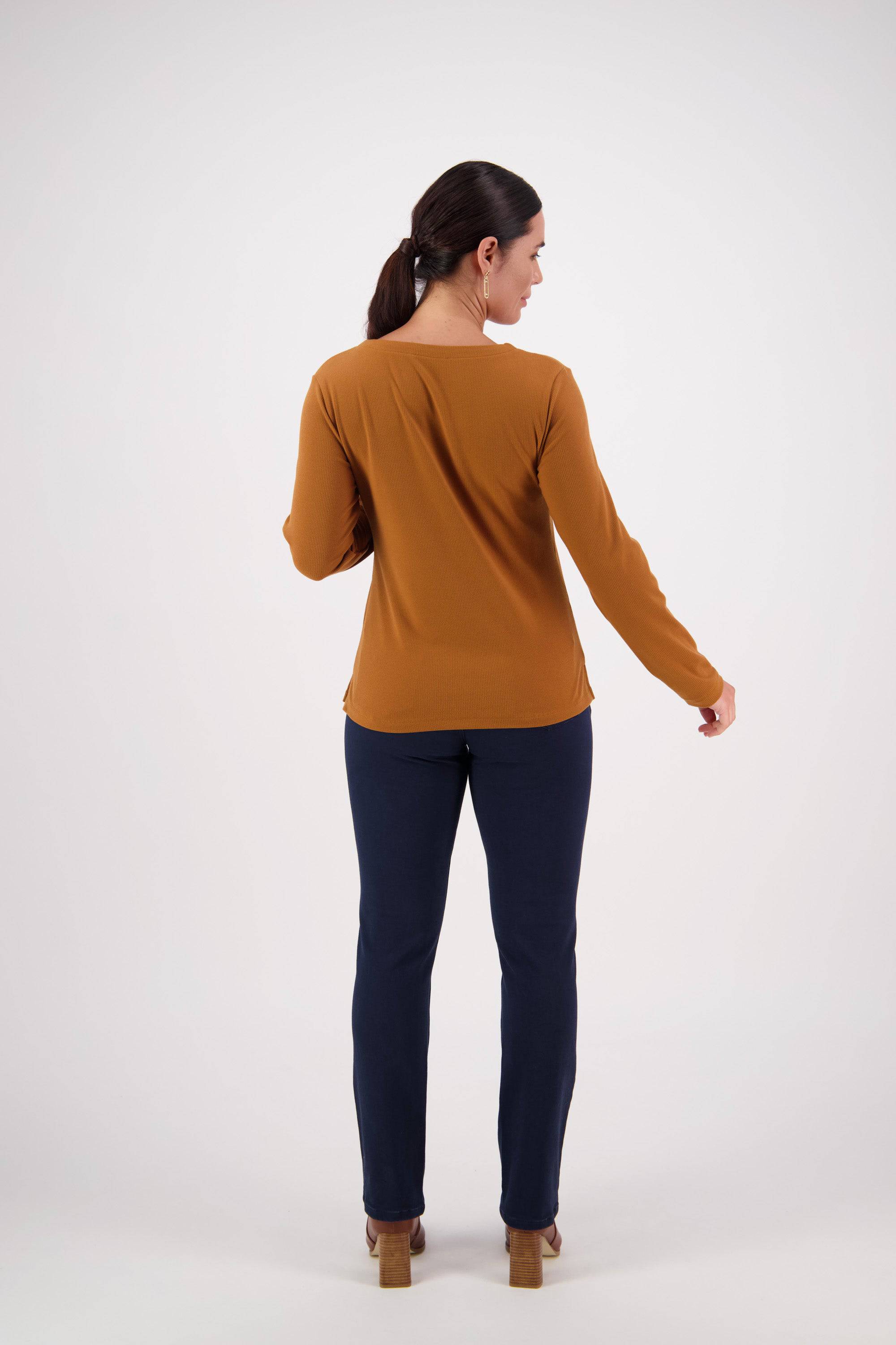 A woman with dark hair in a ponytail stands facing away, wearing a relaxed fit Vassalli V-Neck Top in Tobacco made from soft rib fabric, paired with dark blue pants and heels, against a plain white background.
